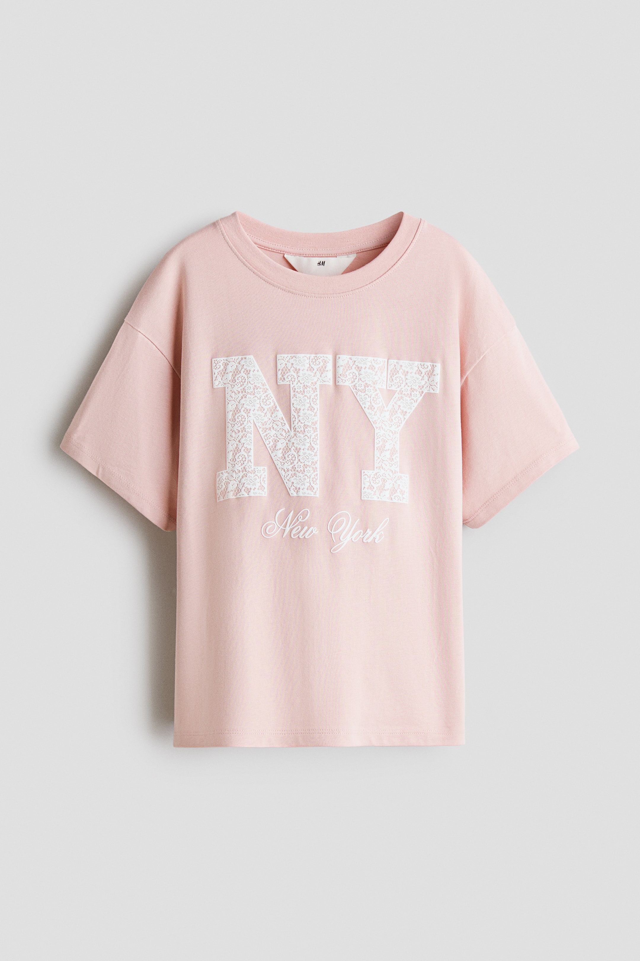 Grösseres Bild ansehen: Ein rosafarbenes T-Shirt zeigt die Vorderseite und verfügt über einen Rundhalsausschnitt sowie kurze, überschnittene Ärmel. Auf der Vorderseite ist in grosser weisser Schrift ein „NY“-Schriftzug mit Spitzenmuster zu sehen, darunter steht „New York“ in Schreibschrift.
