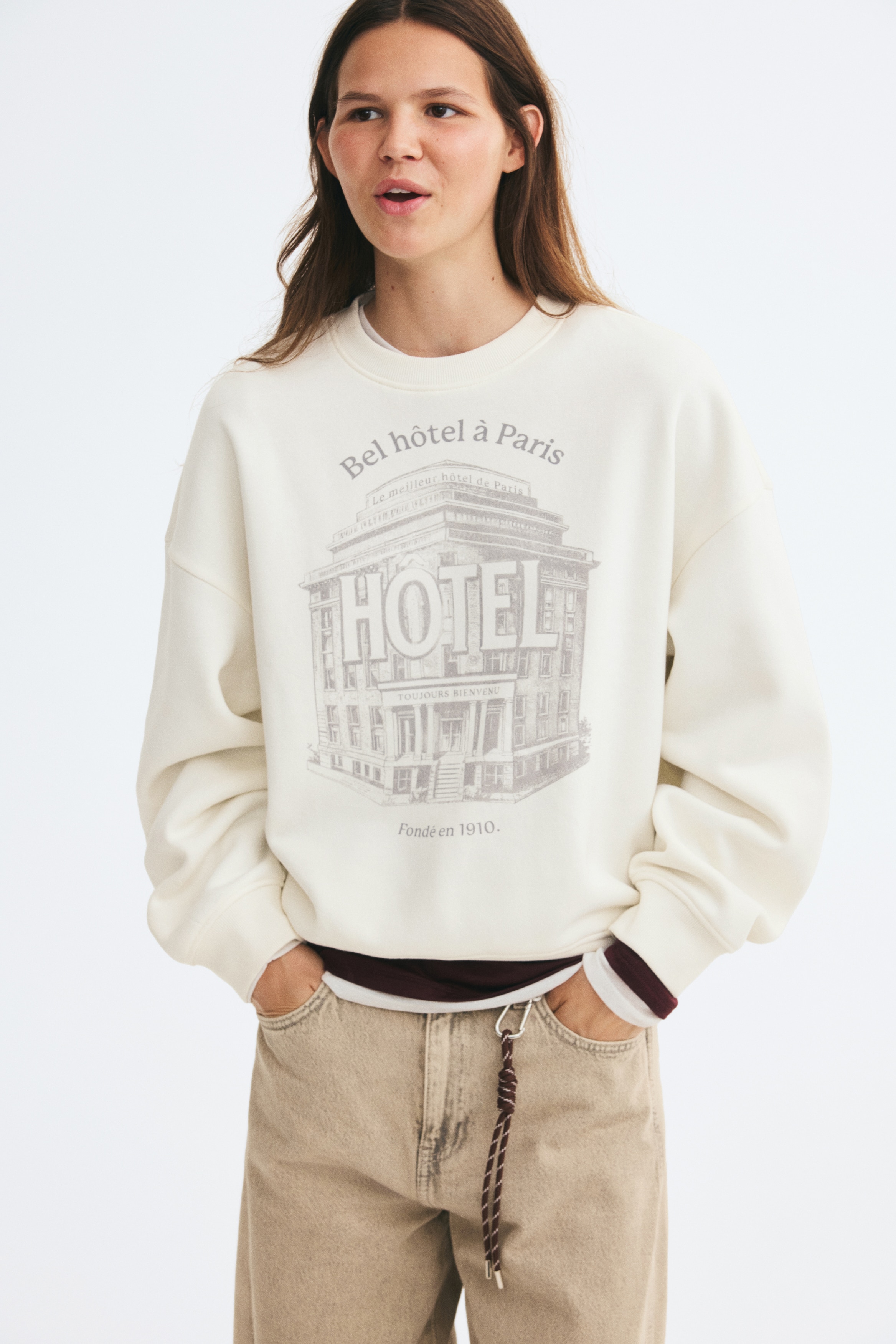 An off-white, oversized sweatshirt is shown from the front, featuring a ribbed crew neck, dropped shoulders, and ribbed cuffs and hem. The center front displays a grey graphic print depicting a classic hotel building with the text "Bel hÃ´tel Ã  Paris" and "FondÃ© en 1910."