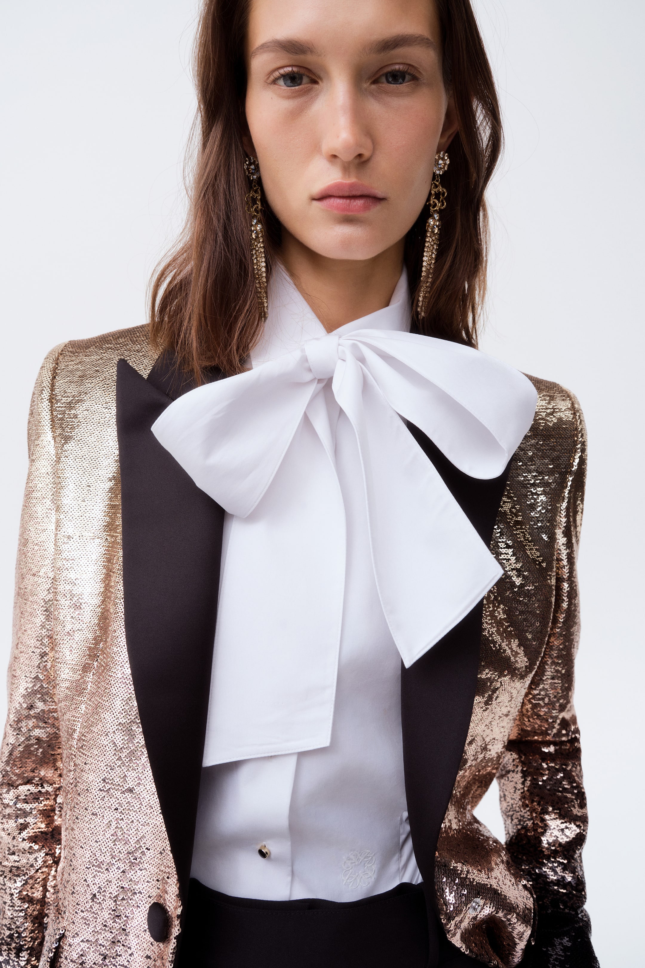 View larger image: A sequined blazer in a metallic rose-gold hue is shown front-facing, featuring contrasting black peak lapels and structured shoulders. The blazer is worn over a white collared shirt with a large white bow tie, showcasing its tailored silhouette and formal styling.