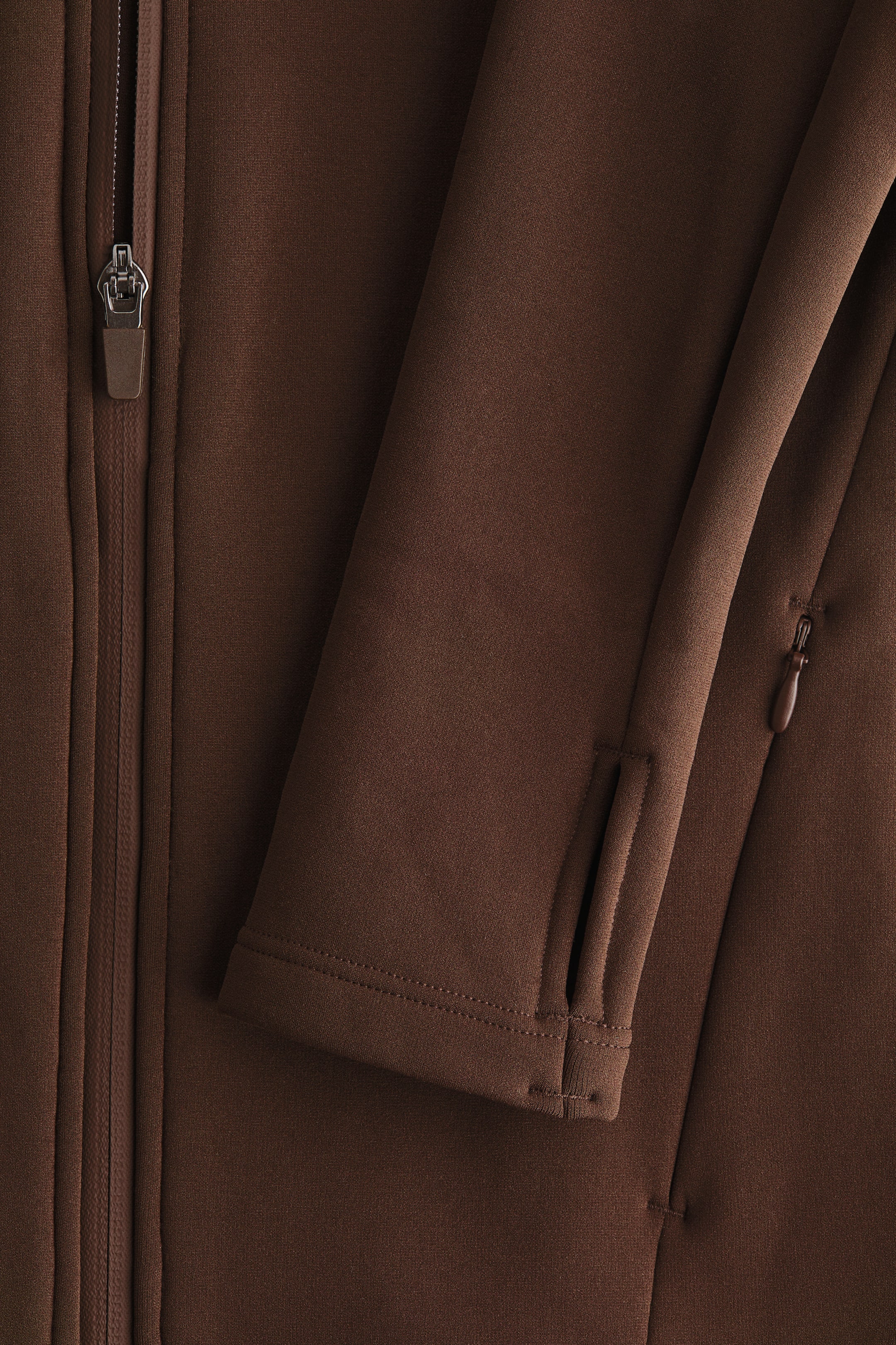 Ampliar la imagen: El producto es una prenda marrón, mostrada desde el frente, con un tirador de cremallera plateado con una lengüeta marrón en una cremallera. Una manga se extiende hacia la derecha, mostrando un puño con un agujero para el pulgar y un pequeño bolsillo con cremallera más abajo en la prenda.