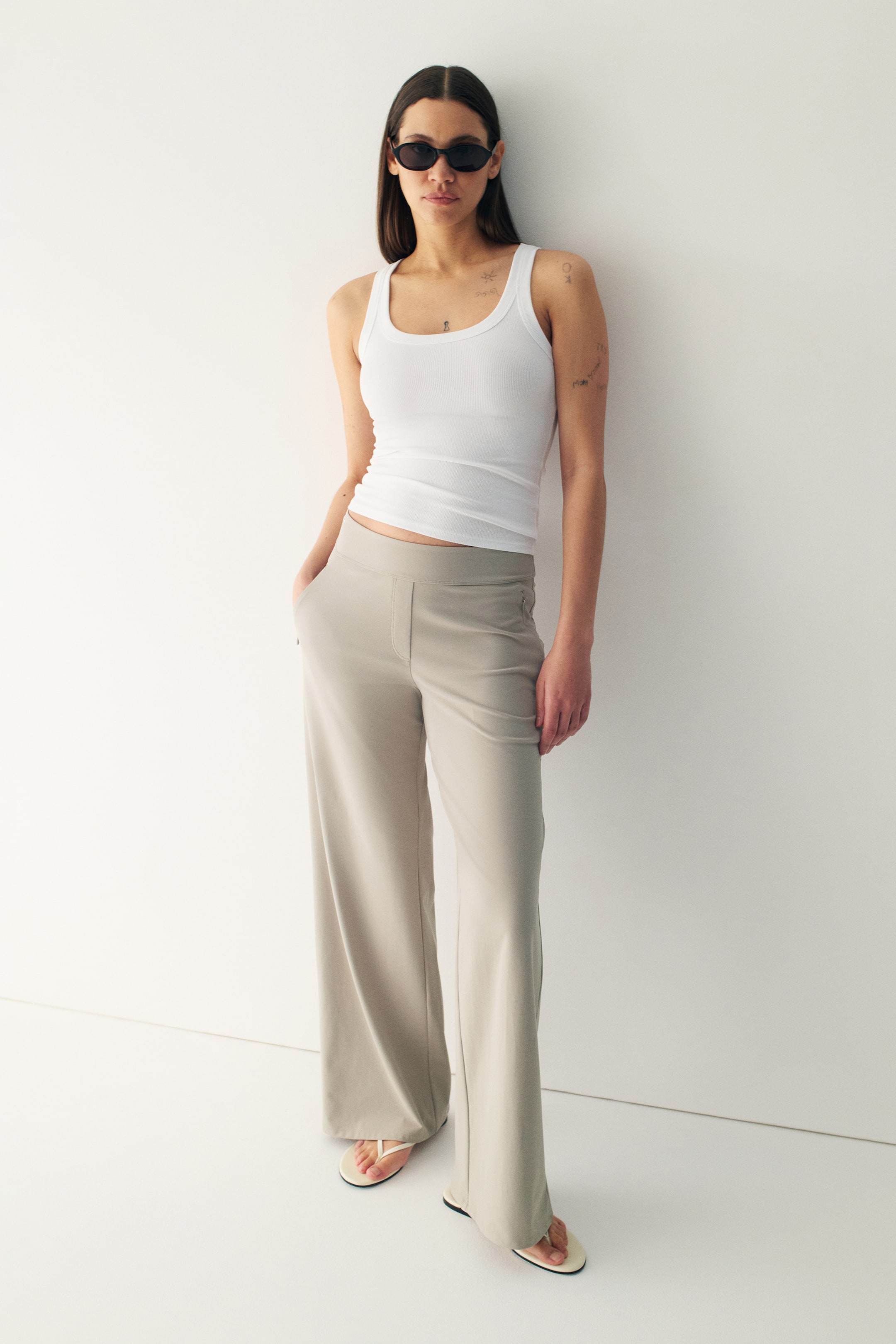 View larger image: The model is showcasing light beige trousers with a relaxed fit and wide legs. These trousers come with an elasticated waistband, a stitched fake fly, and decorative front pockets. The model casually rests one hand in a pocket, highlighting the design's comfort and style.