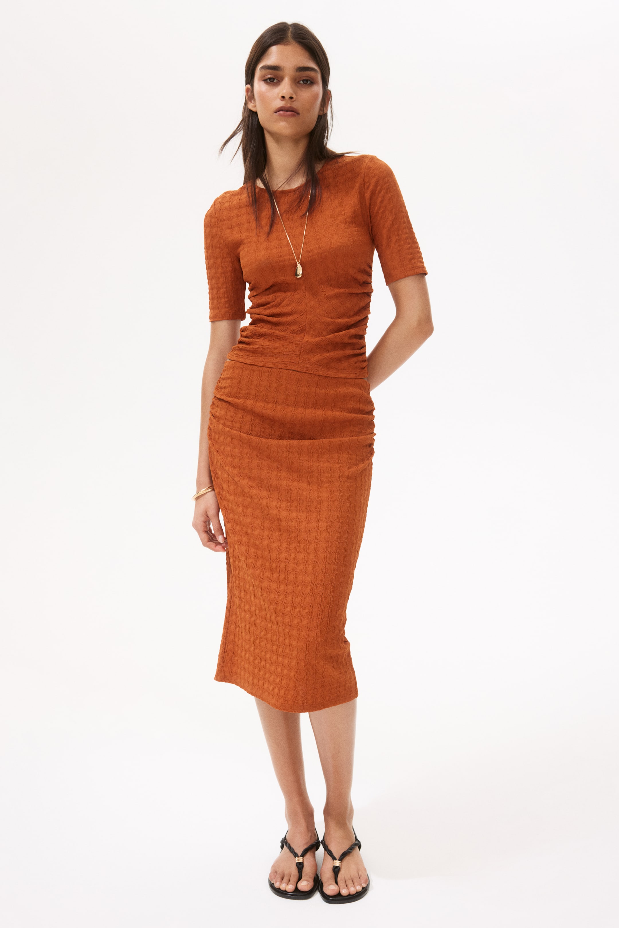 View larger image: A rust orange midi dress featuring a puckered textured fabric, a crew neck, and short sleeves. The dress showcases ruched detailing along the sides of the bodice and skirt, ensuring a close fit to the body.