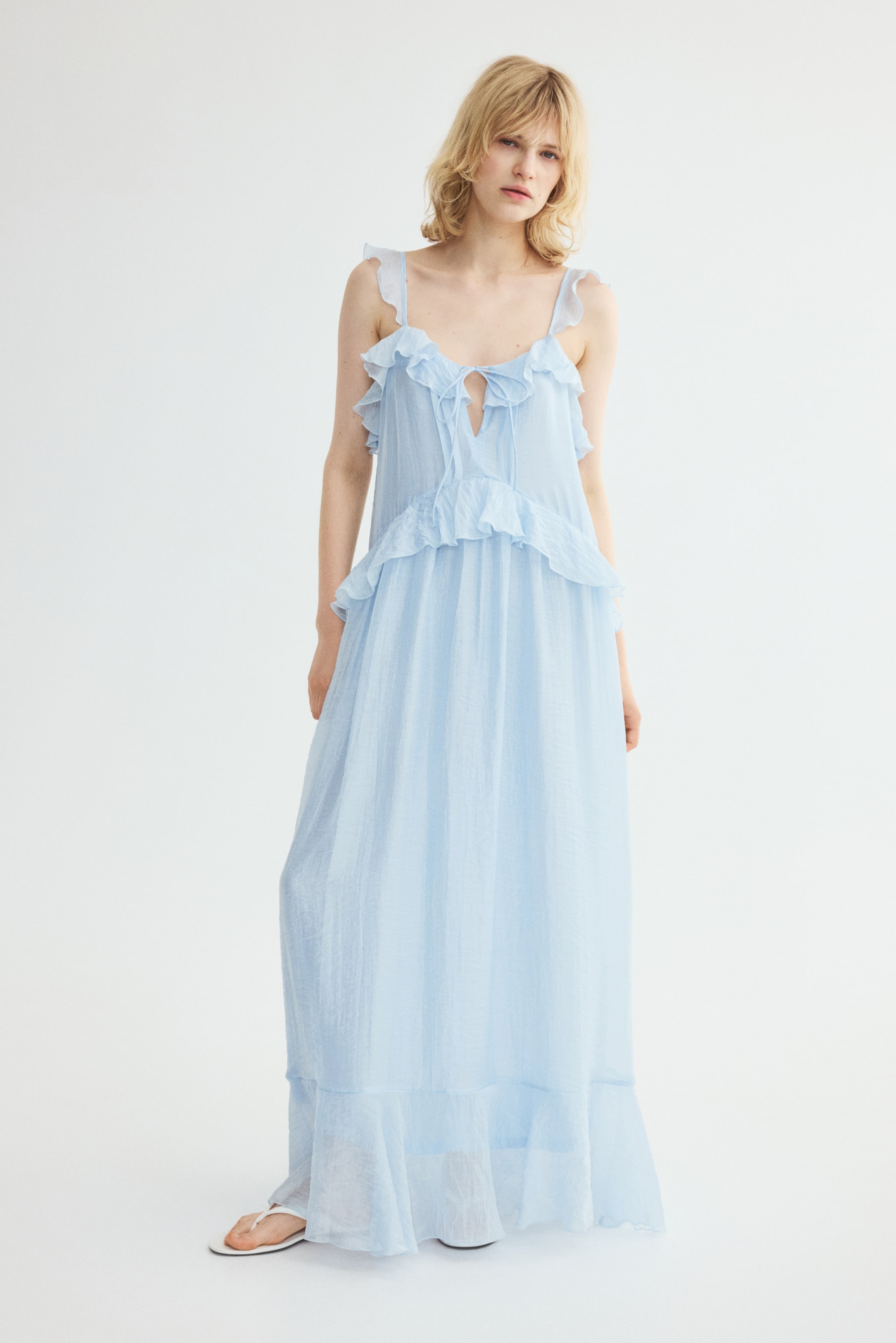Visualizza l'immagine ingrandita: Un abito maxi azzurro, realizzato in un tessuto misto lyocell leggermente stropicciato con una lieve lucentezza, indossato dalla modella, presenta uno scollo quadrato rifinito da volant con apertura a V e lacci, ampie spalline rifinite da volant, pieghe sul corpetto, un taglio in vita arricciato rifinito da volant e una gonna leggermente svasata con un inserto a volant sull’orlo. Indossa anche infradito bianche.
