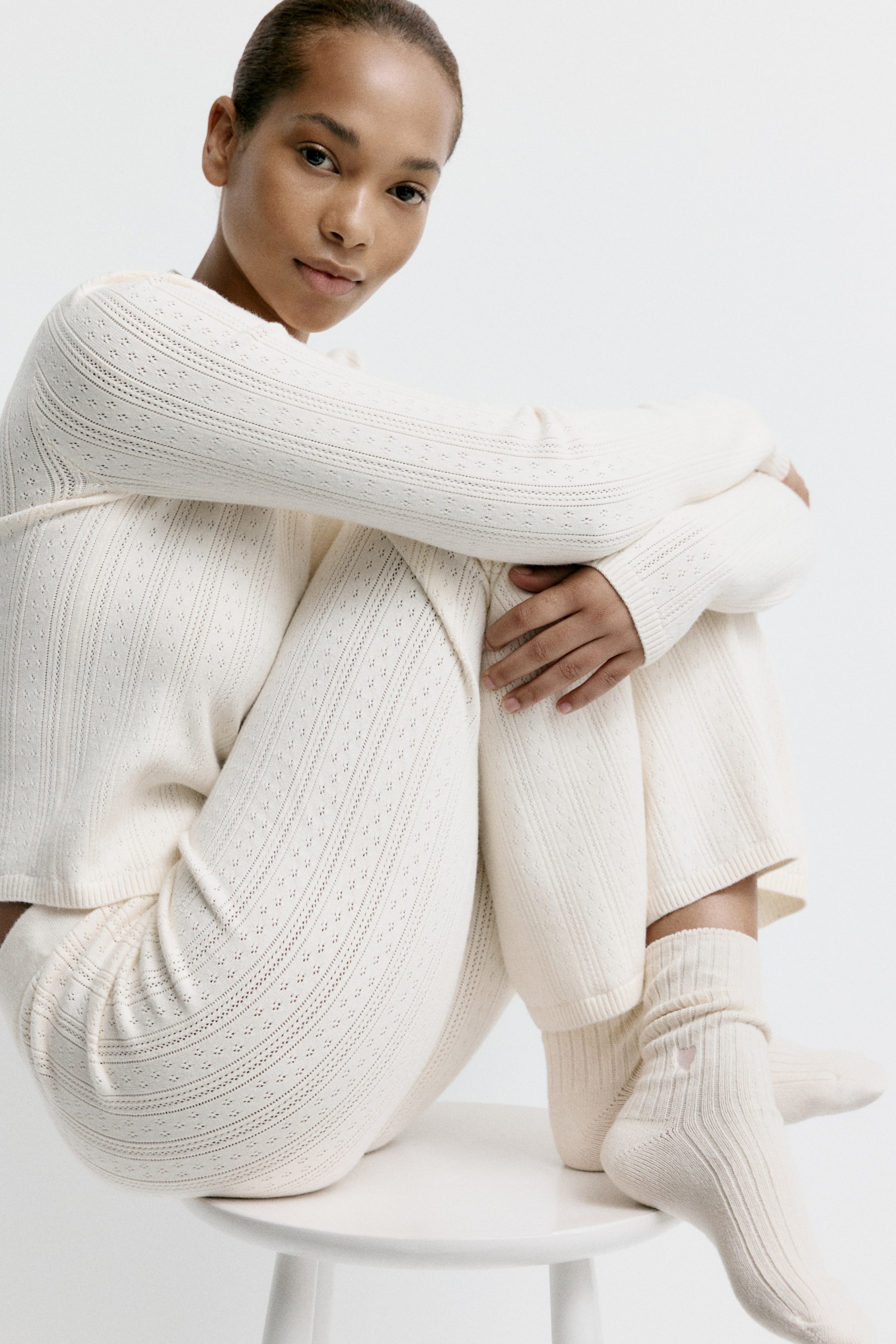 Nagyobb kép megtekintése: Viselésben egy krémszínű, kötött loungewear-szett látható, amely hosszú ujjú felsőből és hozzáillő nadrágból áll. A szettet végigfutó eyelet (lyukacsos) mintázat díszíti, apró, függőleges sorokba rendezett rombuszokkal, míg a megjelenést krémszínű bordázott (ribbed) zoknik egészítik ki, melyeken kis rózsaszín szív hímzés található.