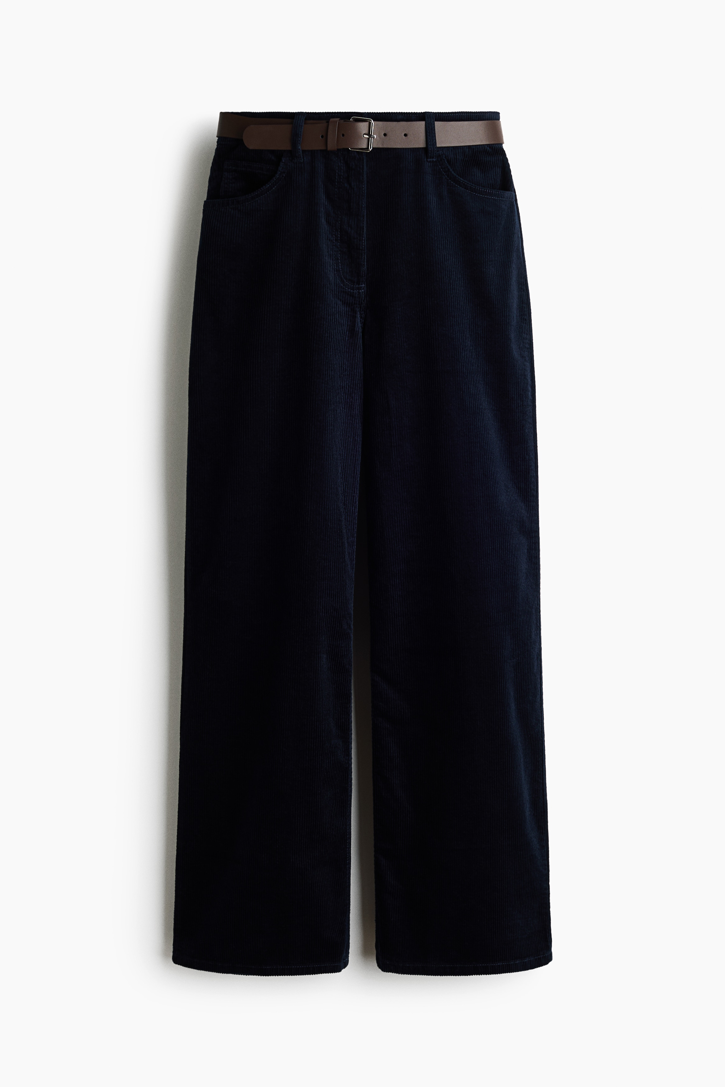 A modell sÃ¶tÃ©t szÃ­nÅ±, magas derekÃº kordnadrÃ¡got visel barna Ã¶vvel, mikÃ¶zben Ã¡llÃ³ pÃ³zban, kezeit az elsÅ zsebekbe helyezve lÃ¡thatÃ³. A nadrÃ¡got sÃ¶tÃ©t denim gombos inggel pÃ¡rosÃ­tottÃ¡k.