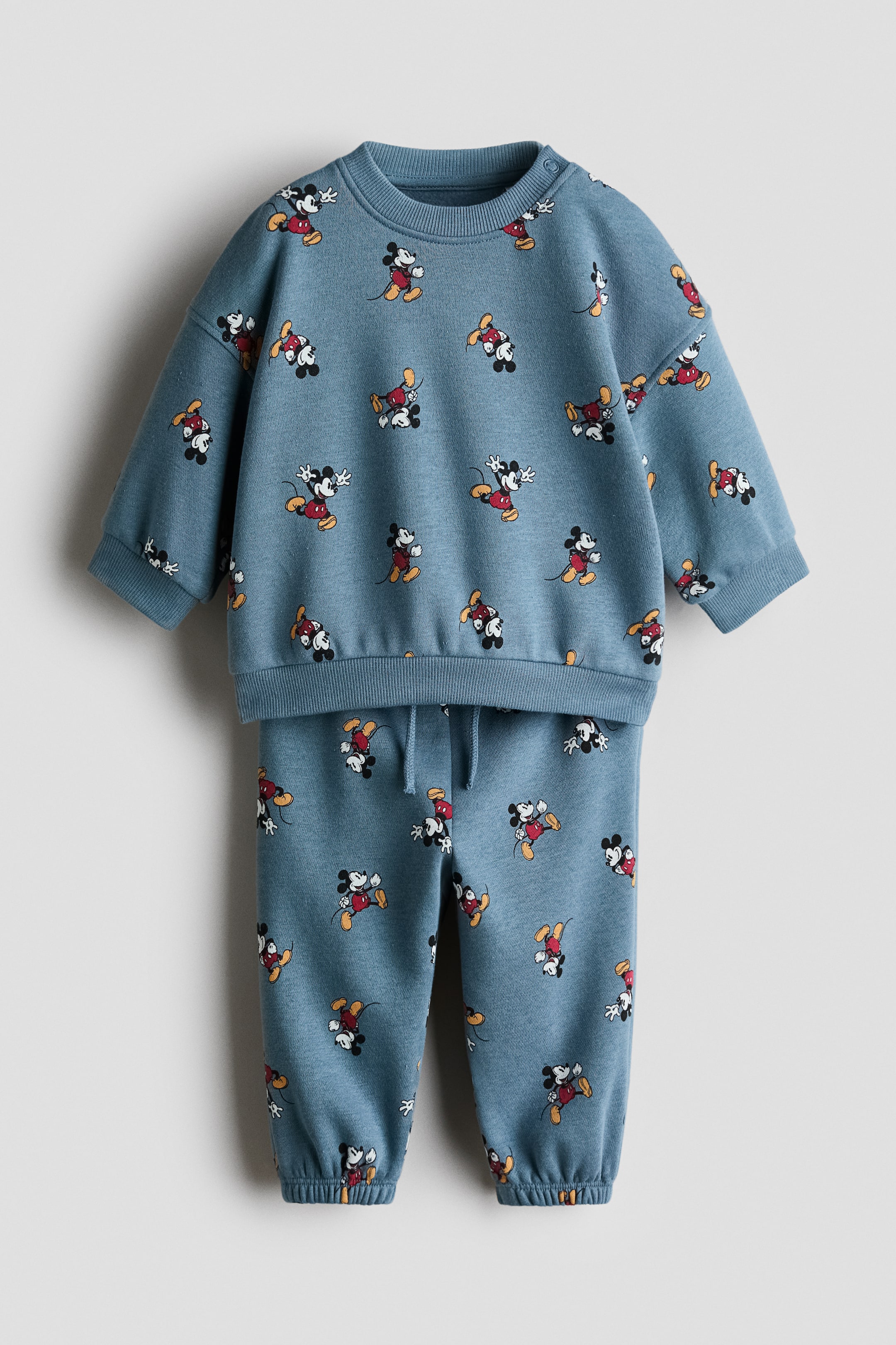 View larger image: This two-piece printed sweatshirt set is displayed from the front, highlighting a blue-grey fabric adorned with an allover Mickey Mouse print. The sweatshirt is designed with a ribbed crew neck featuring snap buttons on the shoulder, ribbed cuffs, and a ribbed hem. Complementing the top, the matching sweatpants come with an elastic drawstring waist and ribbed ankle cuffs.