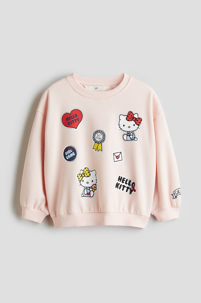 Ein hellrosa Sweatshirt wird von vorne gezeigt, mit einem gerippten Rundhalsausschnitt, langen Ärmeln mit gerippten Bündchen und einem gerippten Saum. Die Vorderseite ist mit mehreren bunten Aufnähern verziert, darunter Hello Kitty-Figuren, ein rotes Herz mit dem Text "HELLO KITTY", ein "BEST FRIEND"-Band und ein "GIRL GANG"-Aufnäher. Ein "GIRL"-Banner-Aufnäher ist ebenfalls auf dem rechten Ärmel sichtbar. - 1