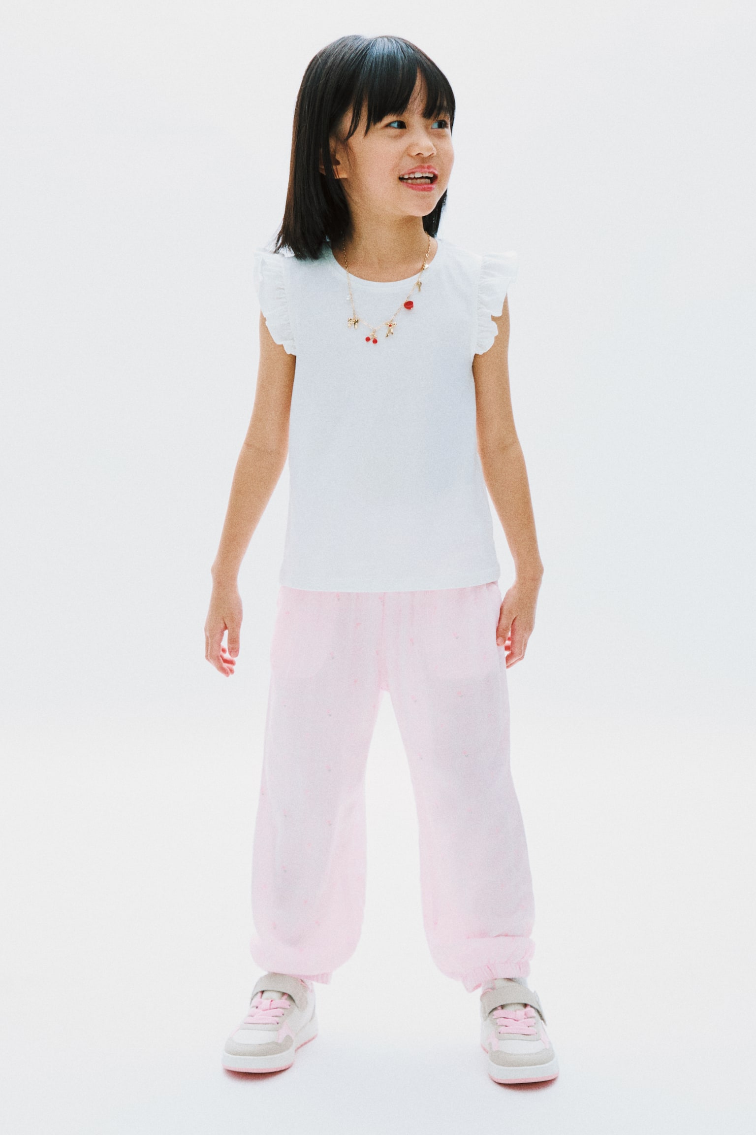 Produk ini, iaitu baju putih tanpa lengan dengan bahu beropol, dipadankan dengan seluar panjang berwarna merah jambu muda yang menampilkan corak berbintik halus dan manset berelastik. Sebuah rantai berwarna emas dengan hiasan loket merah dan emas menghiasi baju tersebut, dan kasut sukan berwarna merah jambu muda, putih, dan kelabu dengan tali velcro melengkapkan pakaian ini. - 1