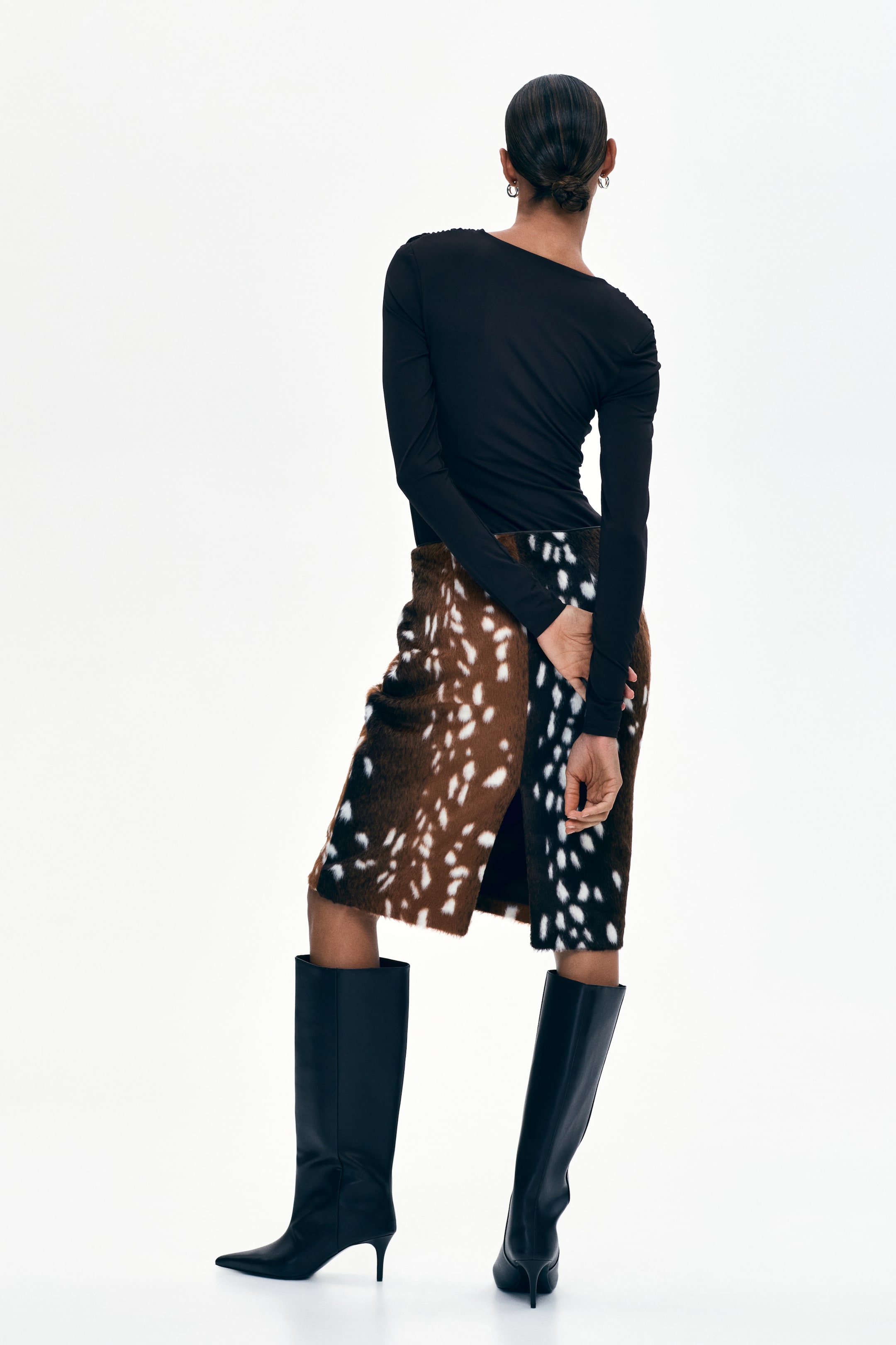 View larger image: The knee-length skirt features a unique split design, with one side showcasing a brown background adorned with irregular white spots, while the other side presents a black background with similar irregular white spots. Viewed from the back-left side, this perspective highlights the distinct pattern and a subtle front slit.