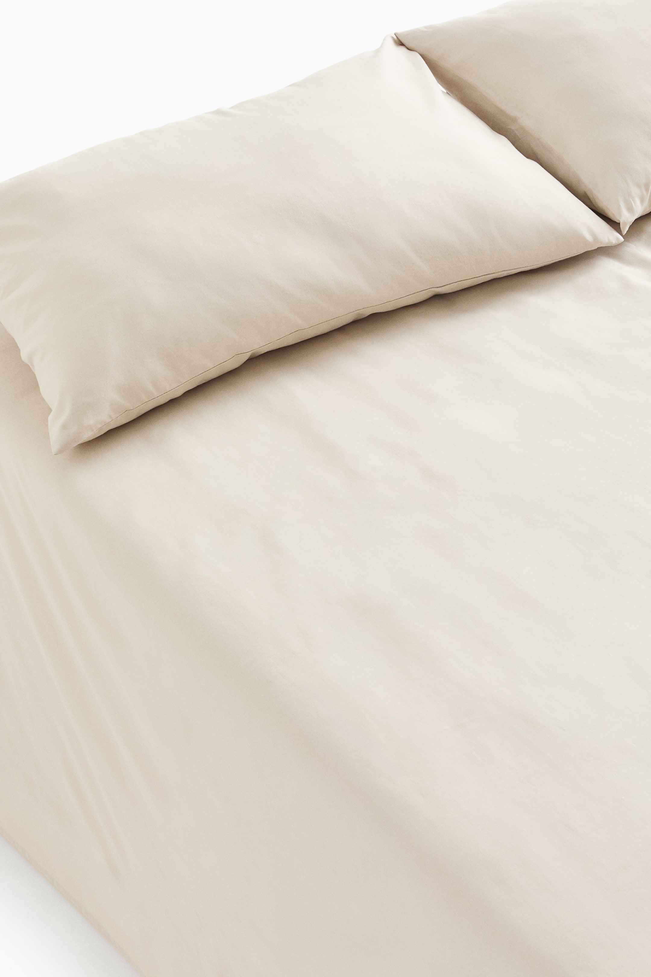 View larger image: A light beige bedsheet set is displayed with a flat sheet spread across a surface, viewed from a slightly elevated angle. Two matching pillowcases are placed side by side at the top, and the soft cotton weave fabric shows subtle natural creases and a smooth texture.
