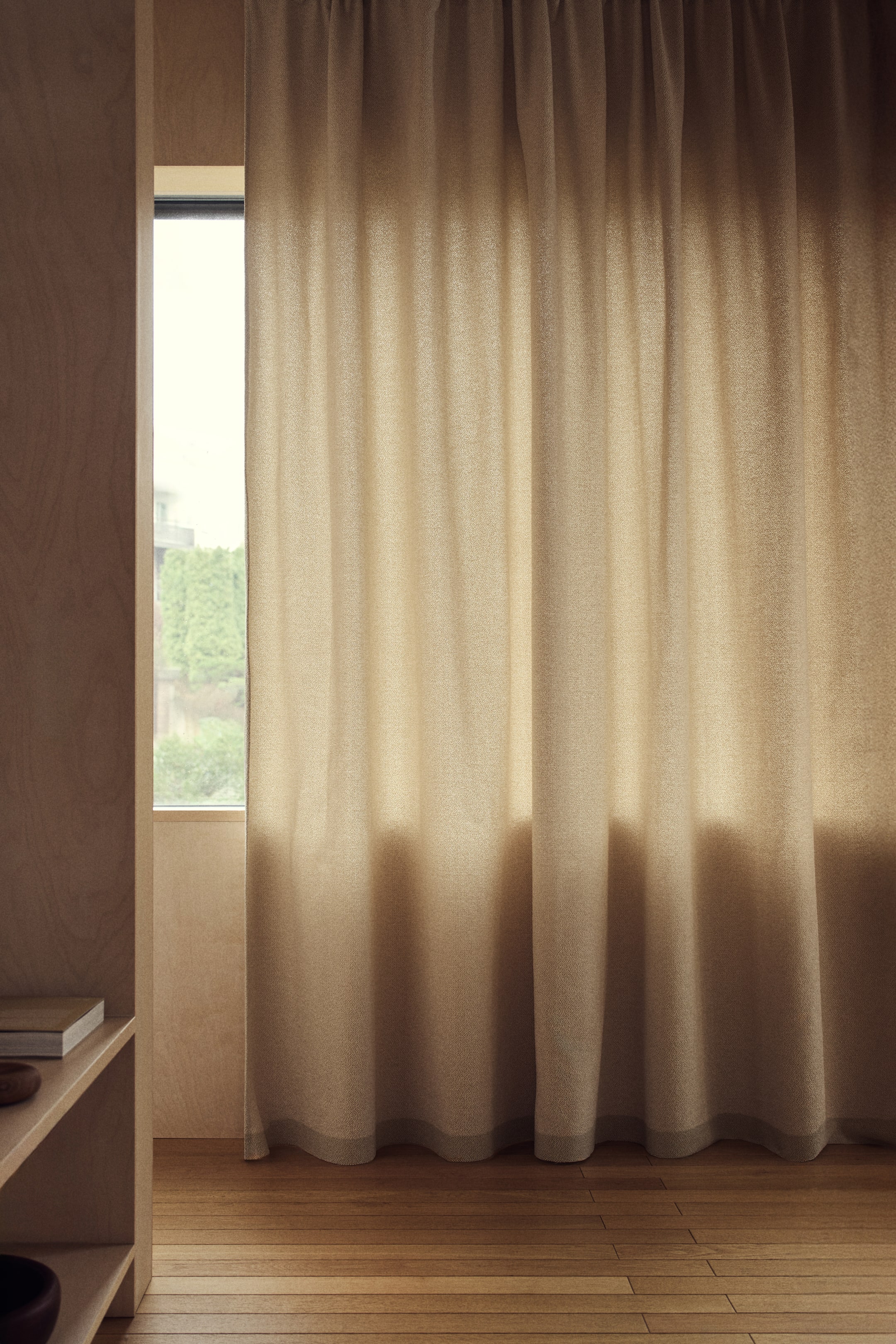 Ampliar la imagen: Una cortina de color beige claro cuelga, extendida a través de una ventana, con suaves pliegues verticales y una tela tejida con textura sutil. Presenta una banda de color sólido más oscuro a lo largo de su dobladillo inferior.