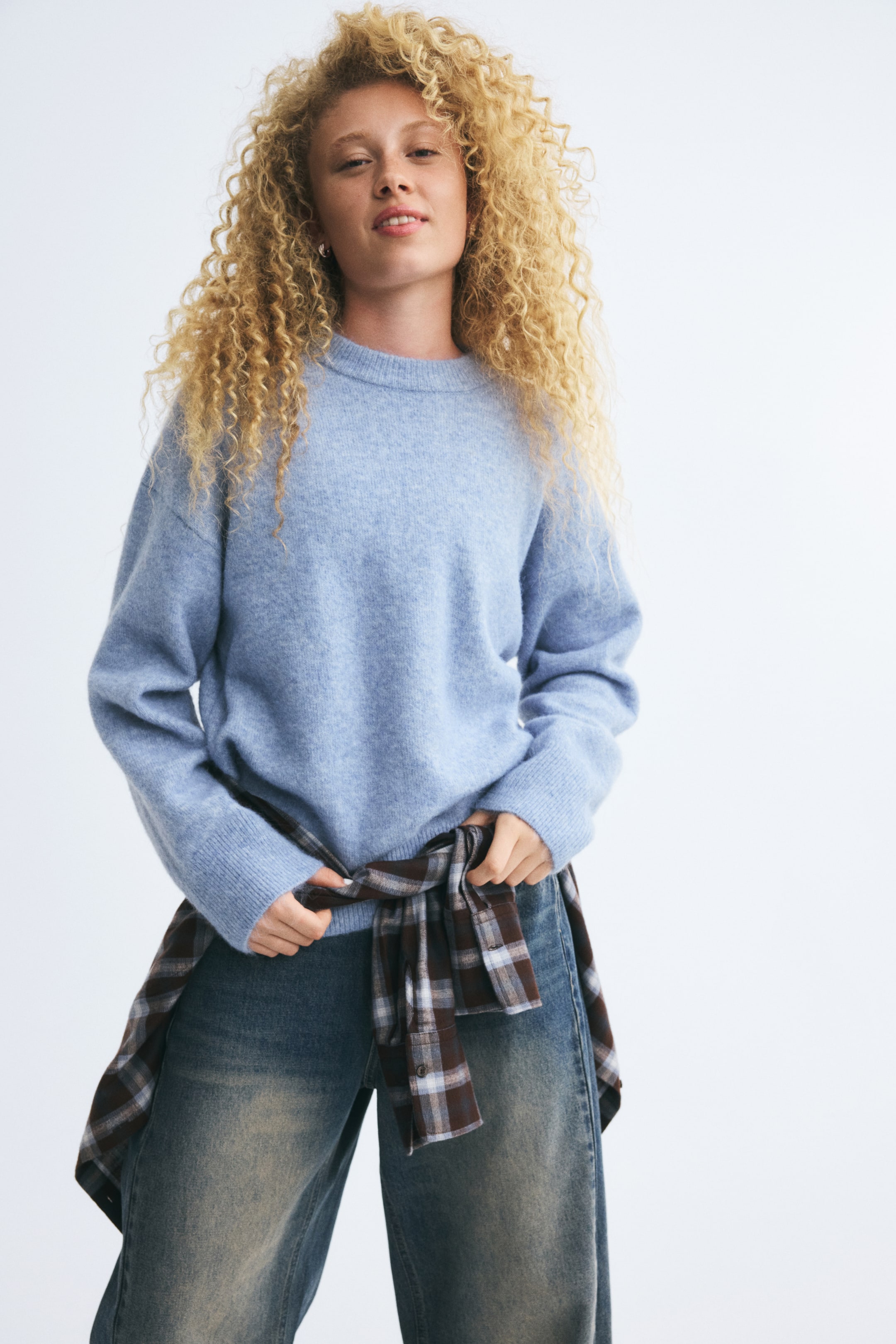 Größeres Bild ansehen: Ein hellblauer, locker geschnittener Pullover aus weichem, gebürstetem Strick wird vom Model getragen und verfügt über einen Rundhalsausschnitt, überschnittene Schultern und Rippbündchen. Das Model hält ein kariertes Hemd, das um die Taille einer ausgewaschenen Wide-Leg-Jeans gebunden ist.