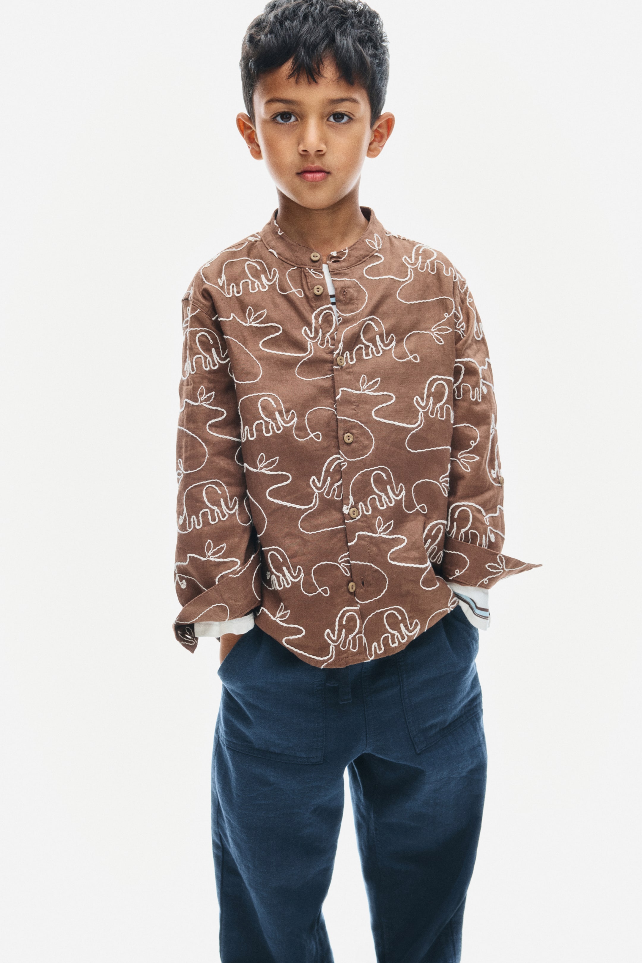 View larger image: A brown grandad shirt featuring an all-over white continuous-line elephant pattern is shown from the front, showcasing a band collar, a button-down front, and long sleeves with rolled-up cuffs.