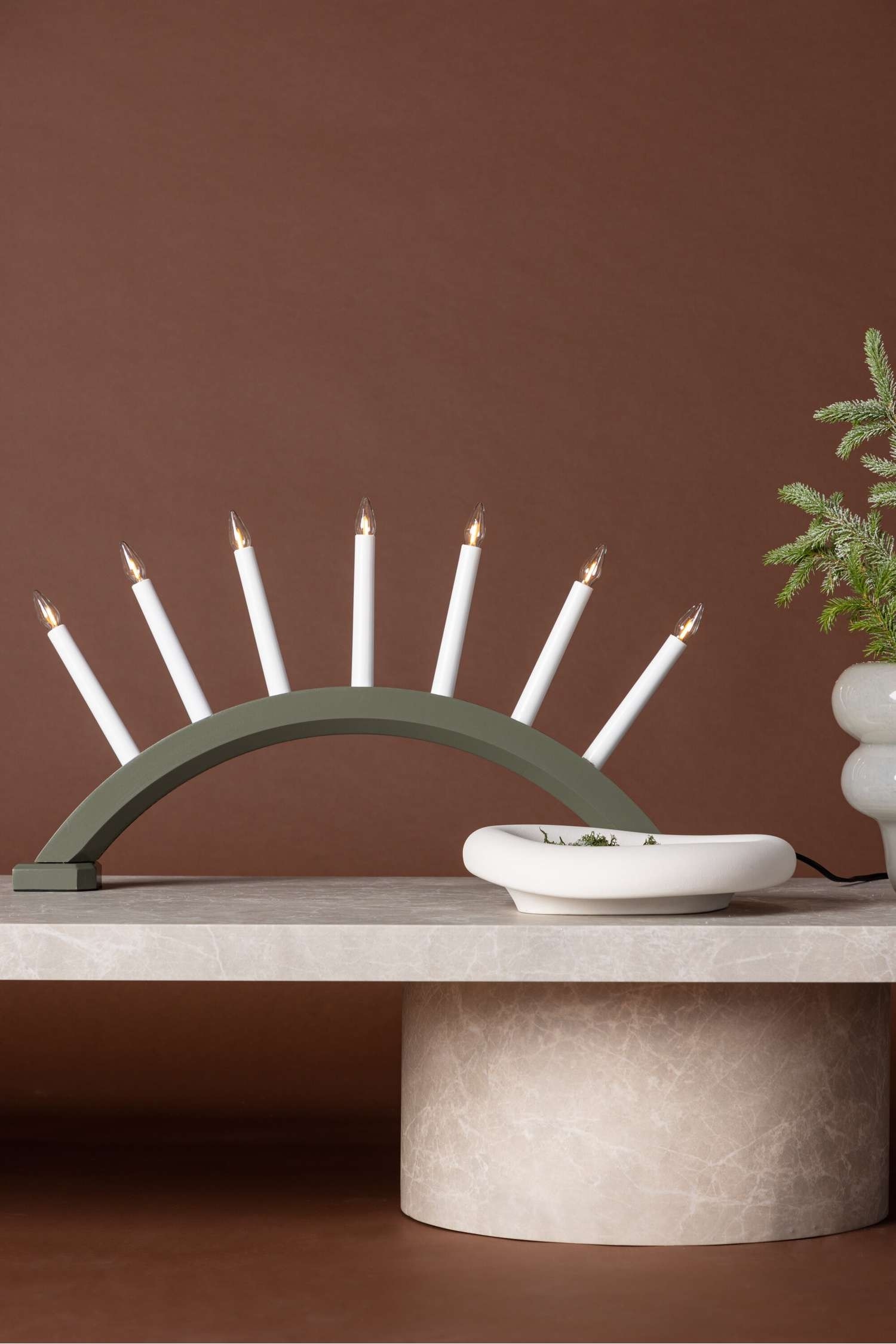 Een tafellamp met een gebogen, saliegroen boogvormig element dat zeven witte kaarsvormige lampen ondersteunt. Een zwarte voedingskabel loopt vanaf de linkerkant van de rechthoekige voet van het armatuur.