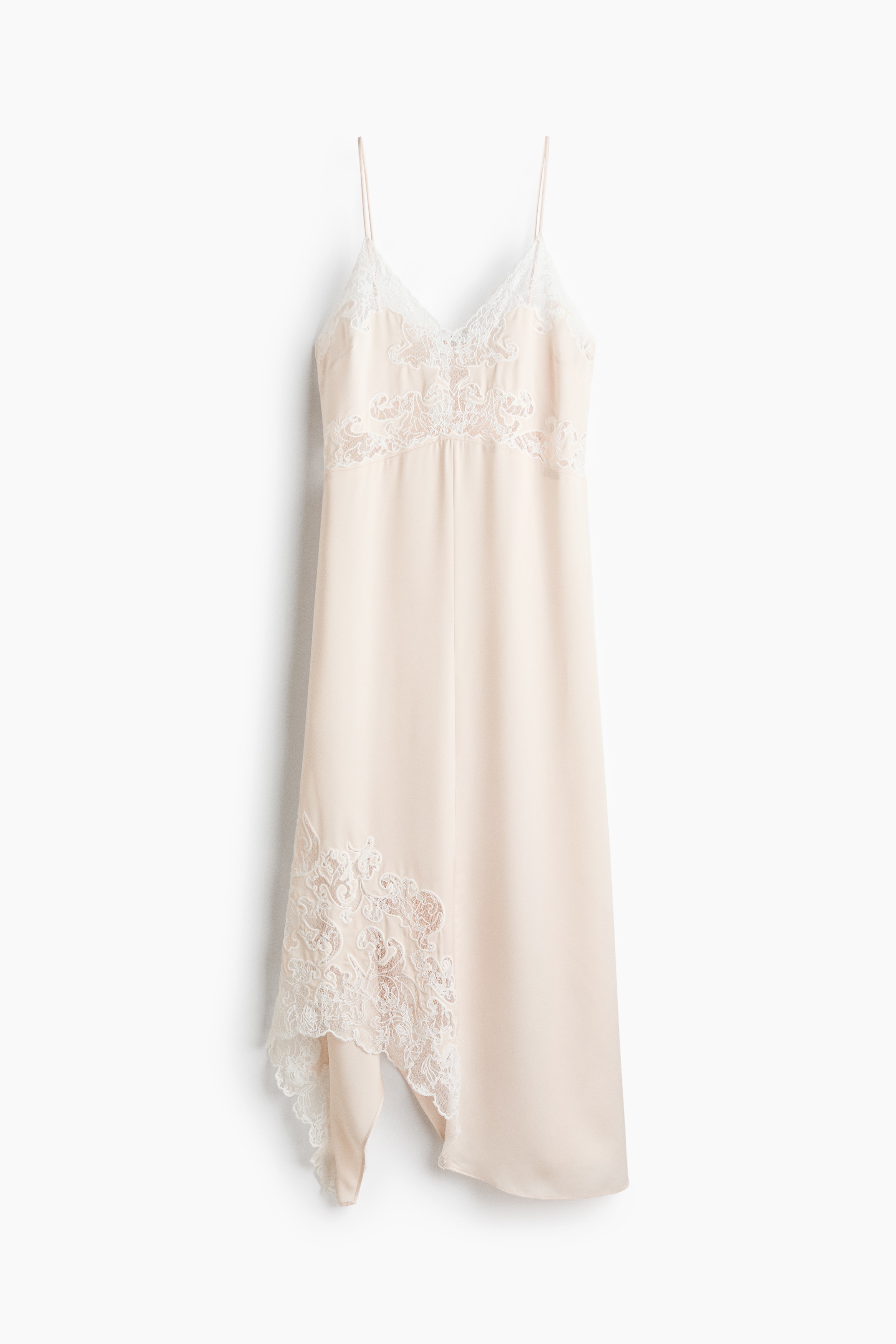 Une longue robe couleur crÃ¨me Ã  fines bretelles spaghetti et Ã  encolure en V est portÃ©e par le mannequin. La robe prÃ©sente de dÃ©licats empiÃ¨cements en dentelle sur le corsage et un ourlet asymÃ©trique en pointe, ornÃ© de dÃ©tails en dentelle assortis, associÃ© Ã  des sandales Ã  talons hauts Ã  brides couleur crÃ¨me.