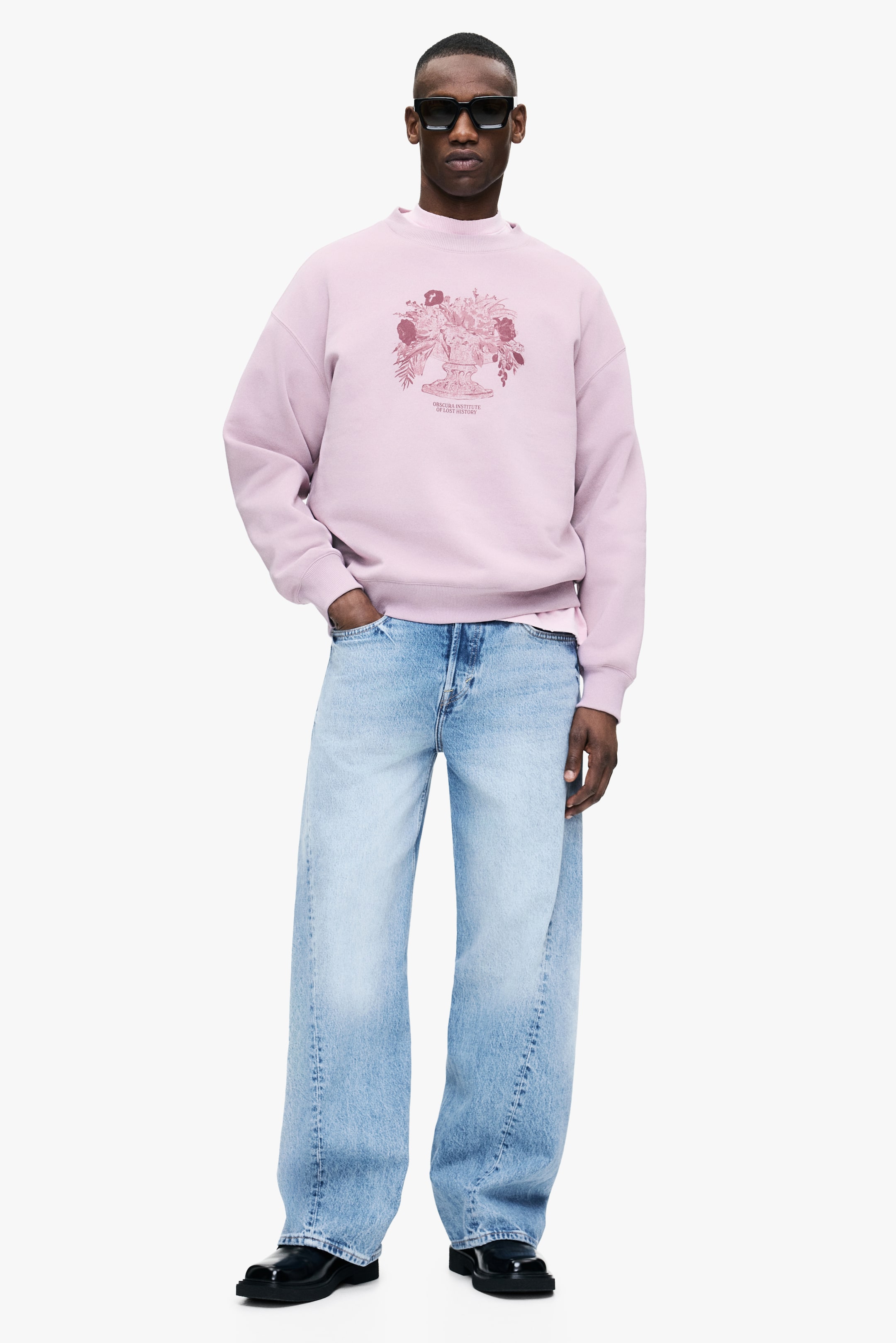 Zobrazit větší obrázek: Růžová mikina s falešným límcem a centrálním grafickým potiskem zobrazujícím květinové aranžmá ve váze s textem je nošena modelkou. Modelka kombinuje mikinu se světle prátými, širokými džínami.