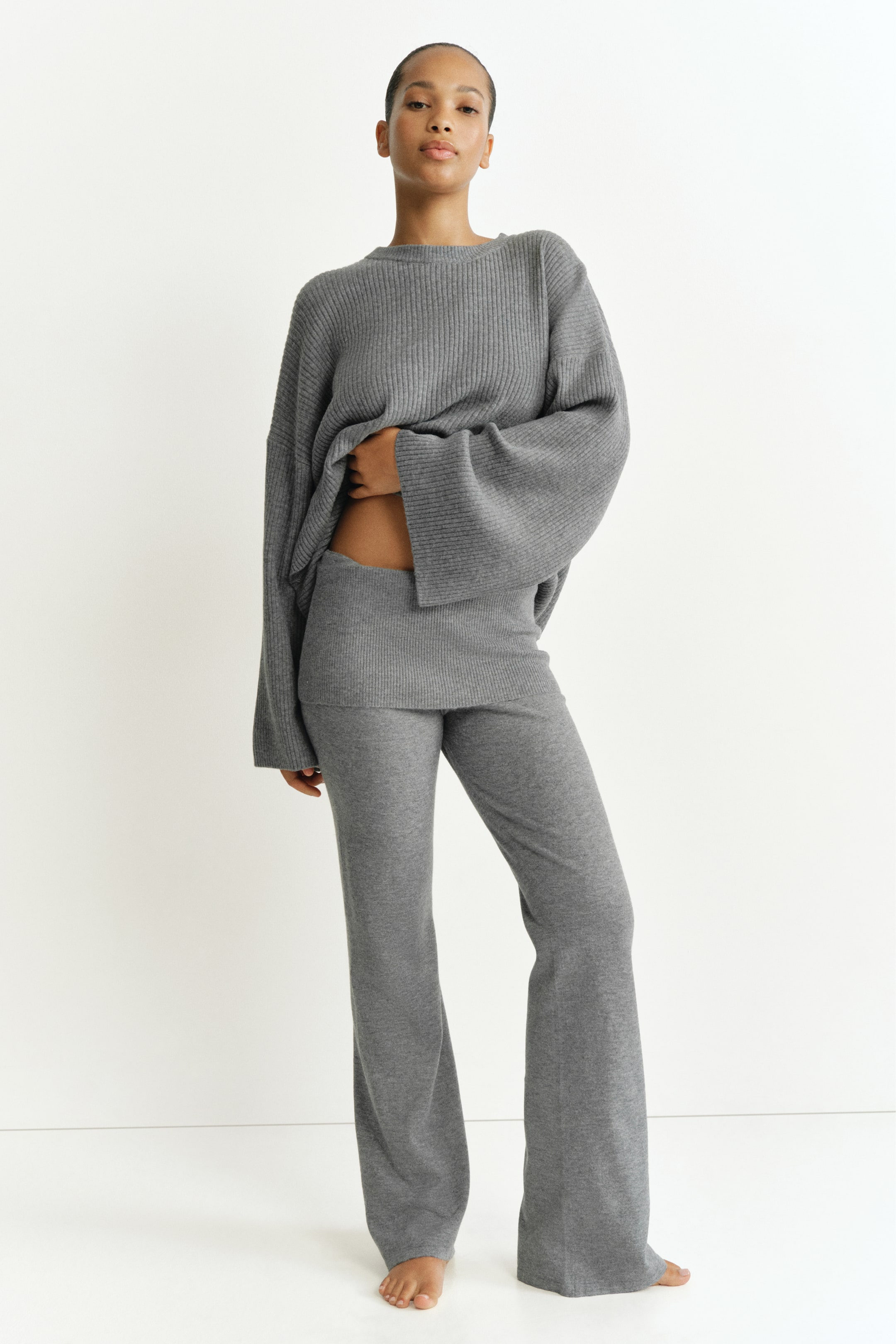 Größeres Bild anzeigen: Eng anliegende Hose aus weichem grauem Strick mit umgeschlagenem Bund und ausgestellten Hosenbeinen, von dem Model getragen. Das Model hebt den Saum eines passenden gerippten Strickpullovers an, um den breiten Bund der Hose zu zeigen.