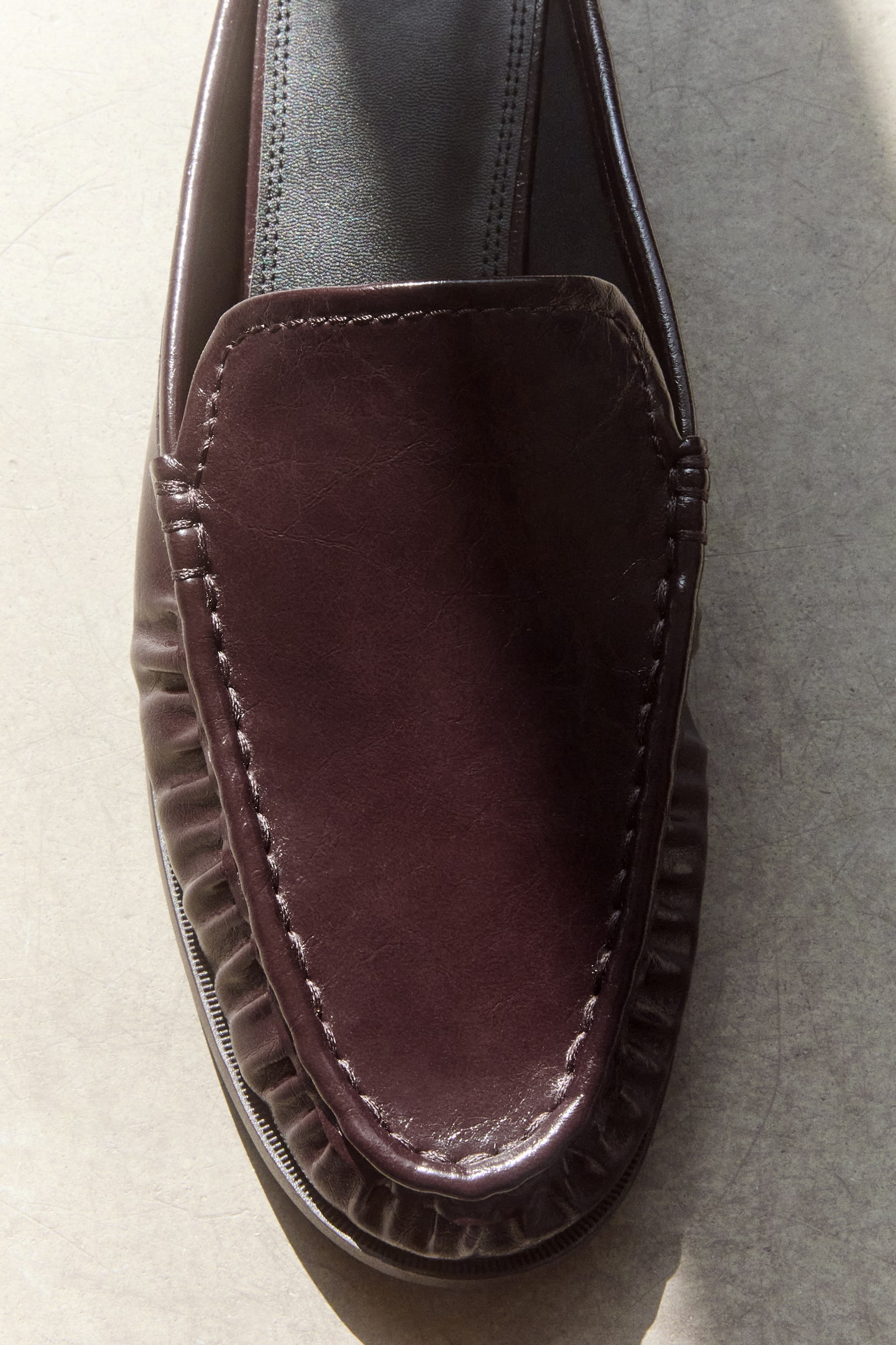 A pair of dark burgundy patent leather loafer mules showcases a high-shine, crinkled texture on the vamp, complete with moccasin-style stitching around the rounded toe, presented side-by-side from a top-down view. - 1