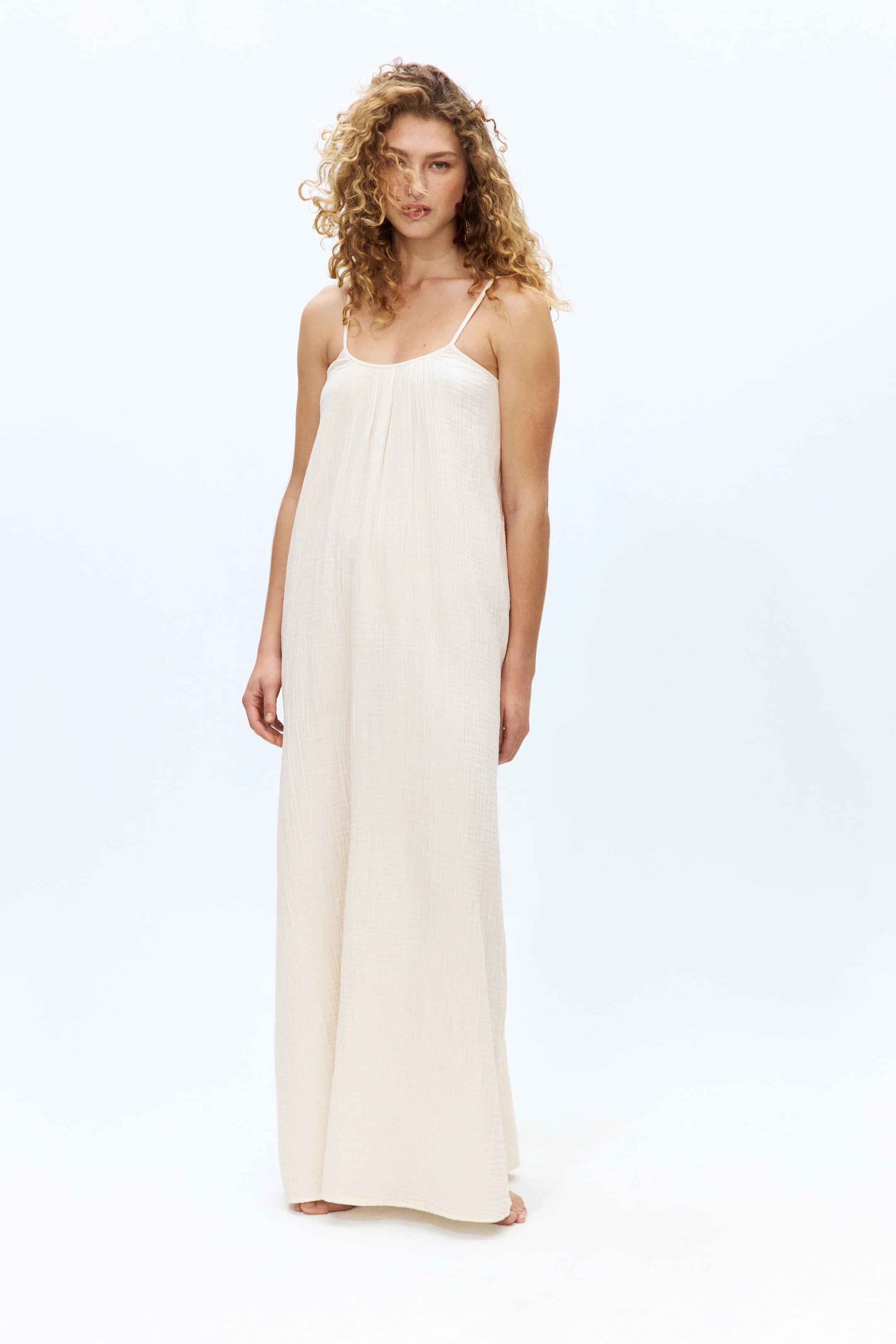 View larger image: A cream-coloured, crinkled gauze wide-leg jumpsuit is being presented from the front. It features thin spaghetti straps, a gathered scoop neckline, and a flowing, loose silhouette.