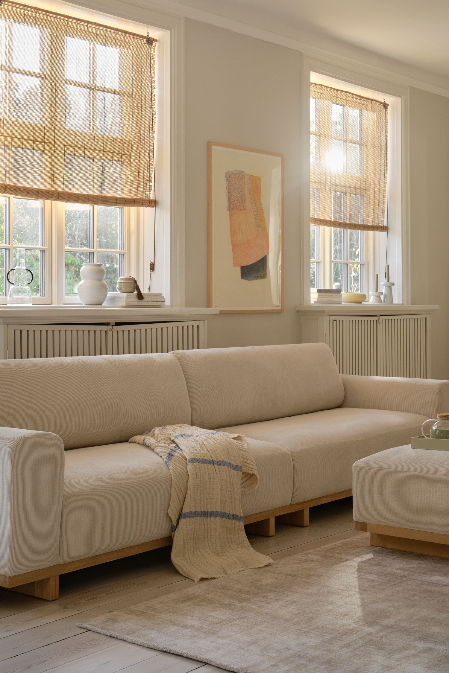 Een laag model bank in crèmekleur staat naar voren gericht en is voorzien van twee grote zitkussens, twee dikke rugkussens en brede armleuningen. Het gestoffeerde frame rust op een lichtgekleurde houten onderstel dat wordt ondersteund door korte, vierkante poten.