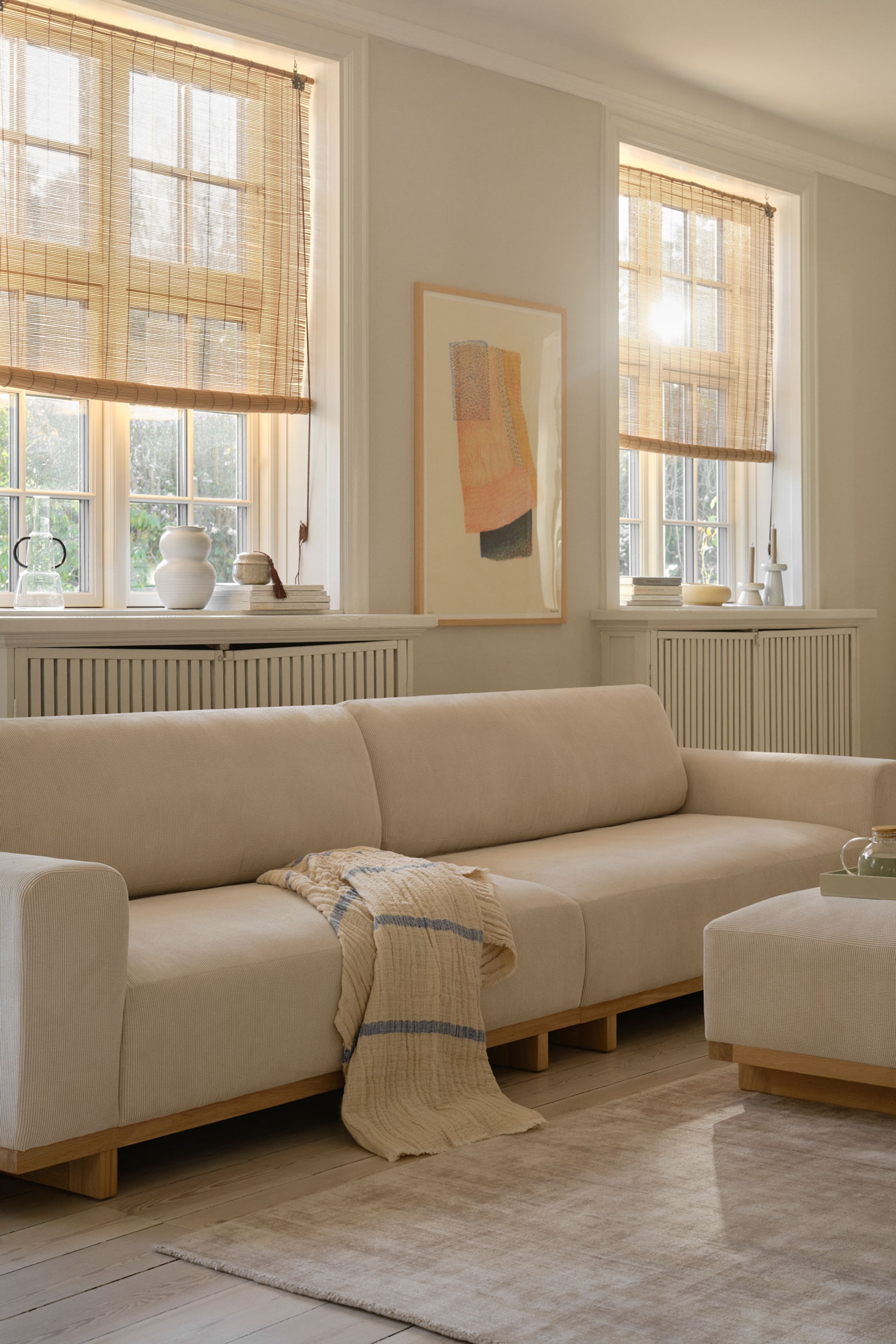 Agrandir l'image: Un canapé trois places au profil bas, doté d’un revêtement en tissu blanc cassé et d’une base plate-forme en bois clair, présenté en vue de face en biais, avec un plaid en maille crème à rayures bleues drapé sur l’assise centrale.
