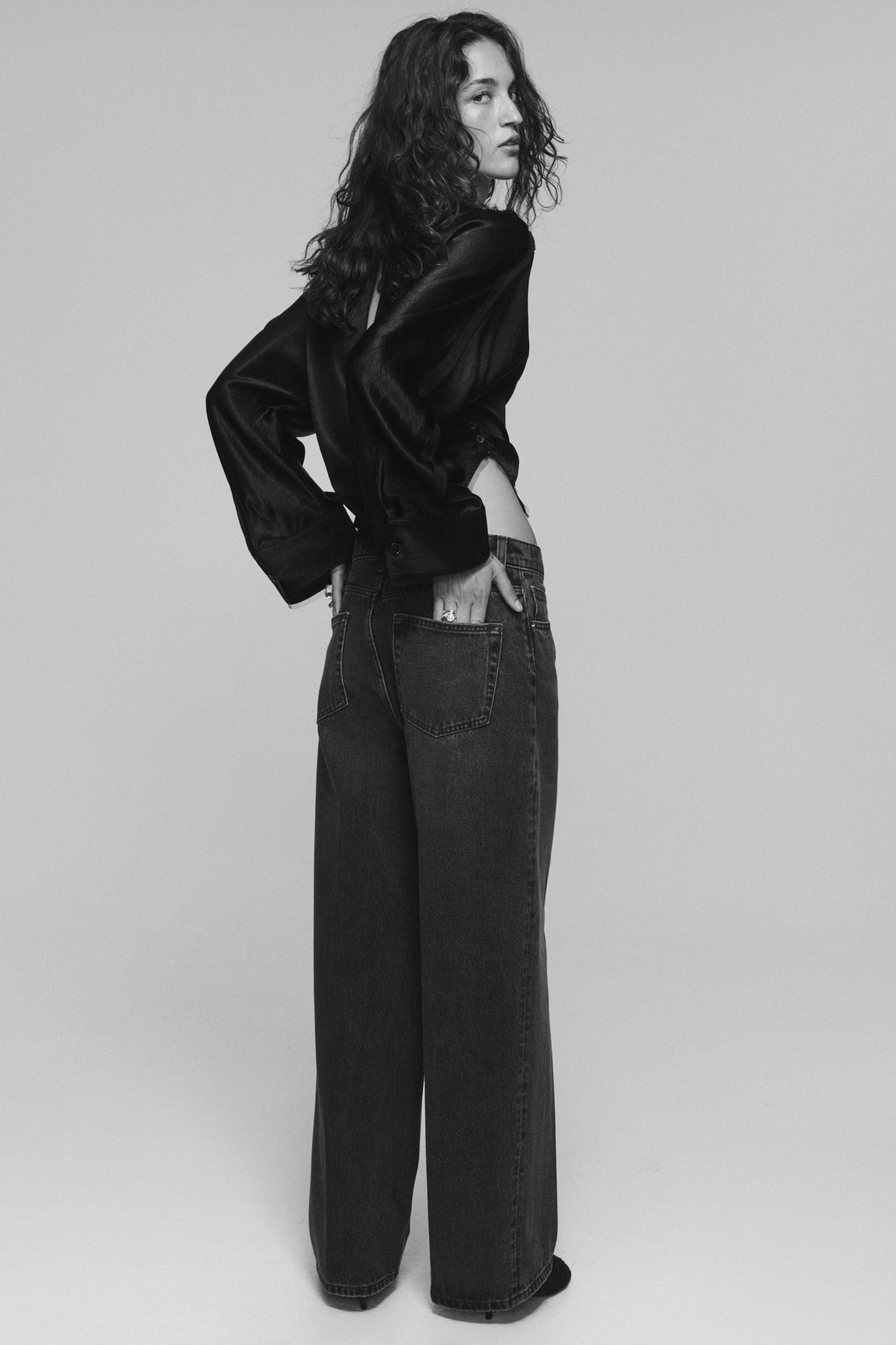 Se større billede: Mørkvaskede denimjeans med brede ben, løs pasform og baglommer bæres af modellen. Modellen står med den ene hånd i en baglomme og styler jeansene med en mørk satinbluse med lange ærmer.