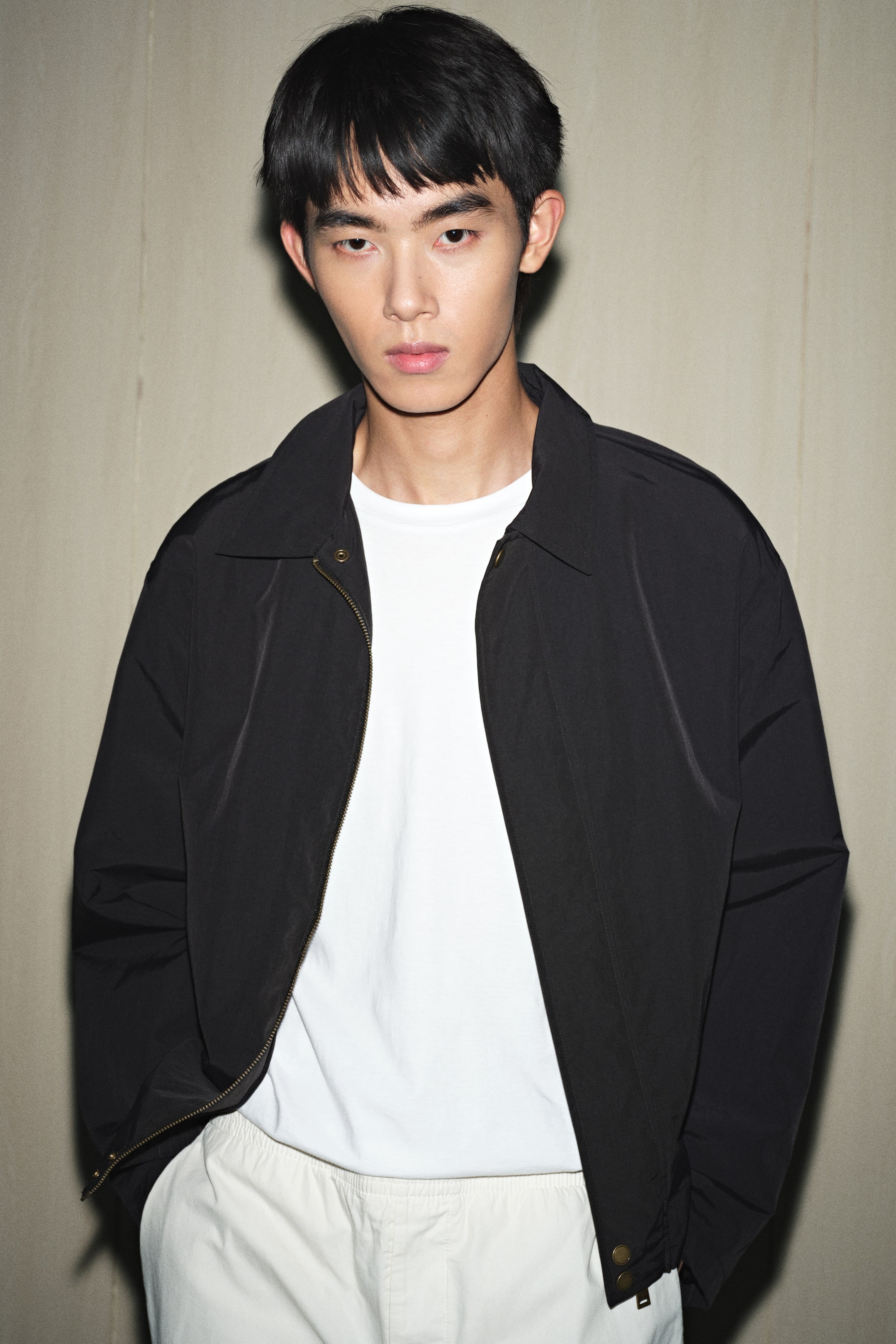 拡大画像を表示: 黒いジャケットは前向きに着用され、白いTシャツの上にジッパーが開いた状態です。それは、尖った襟、前面に真鍮色のジッパー、そして下部の裾に見えるスナップ留め具が特徴です。