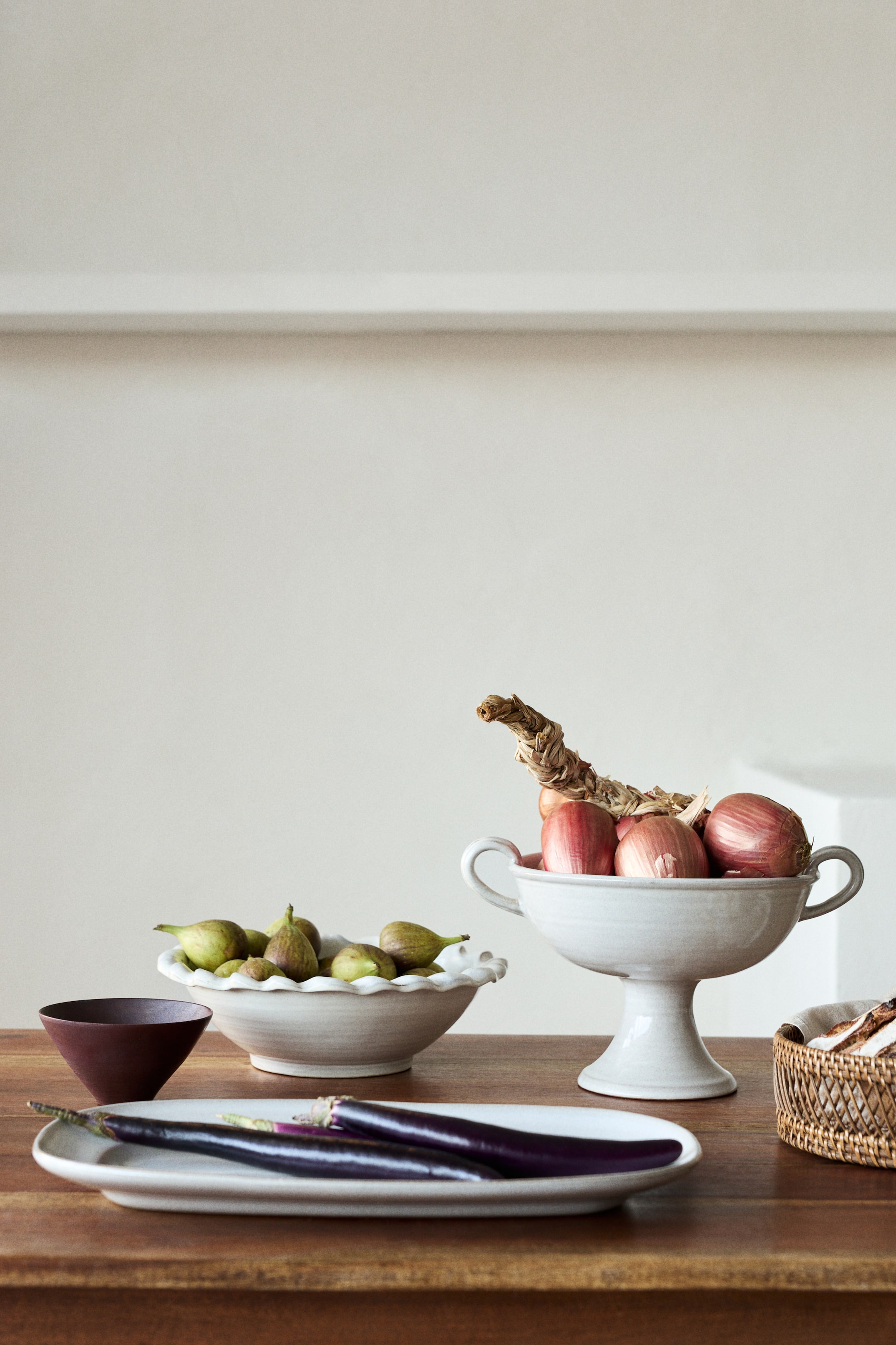 Grotere afbeelding bekijken: Een lichtgrijze keramische kom op voet houdt rode uien vast, met twee kleine handvatten op de rand en een uitlopende voet.