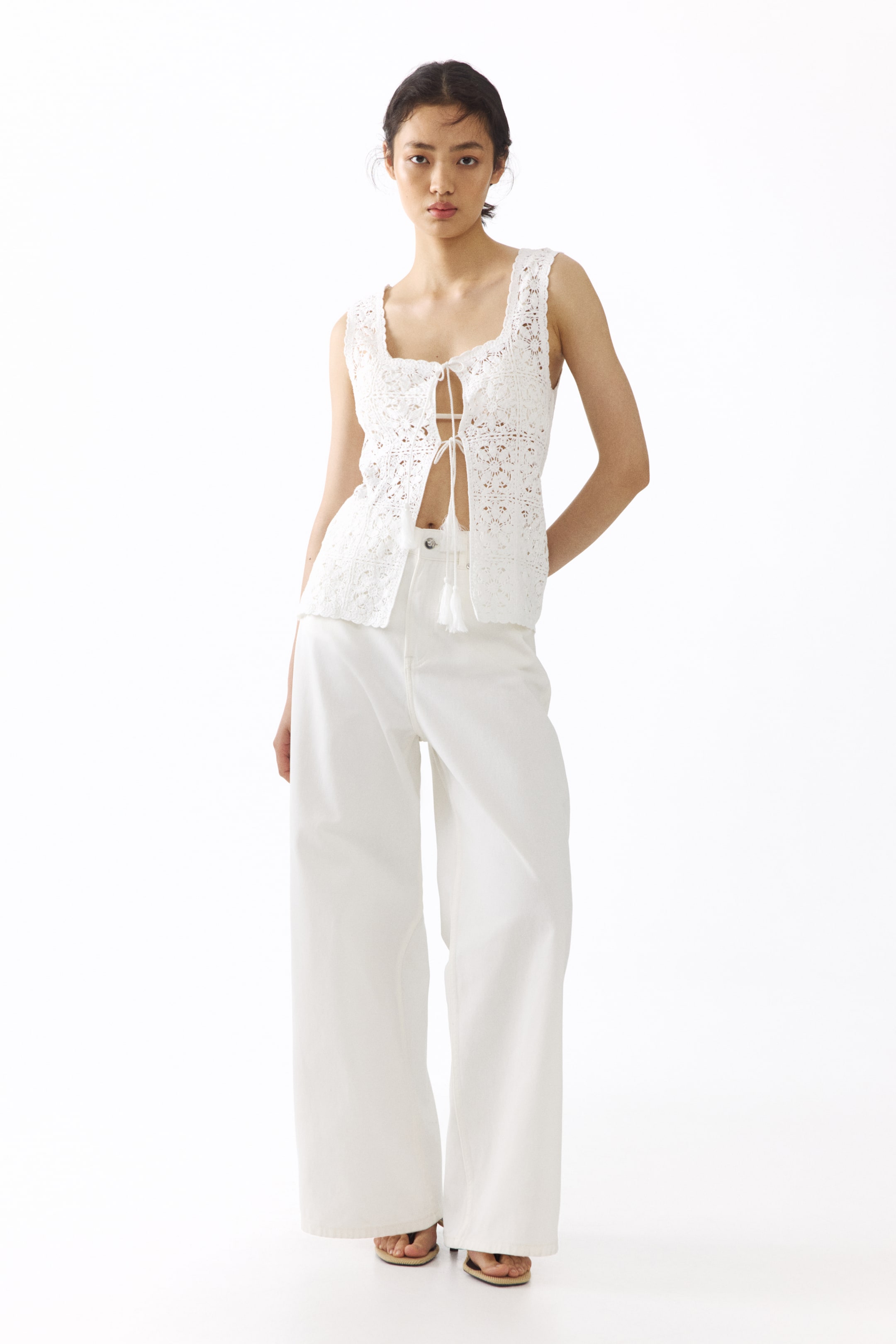 View larger image: The model is wearing a white, fitted, sleeveless pointelle-knit top with a square neckline, scalloped edges, and an open front secured by two pairs of narrow ties with tasseled ends. This top is styled with white wide-leg trousers.