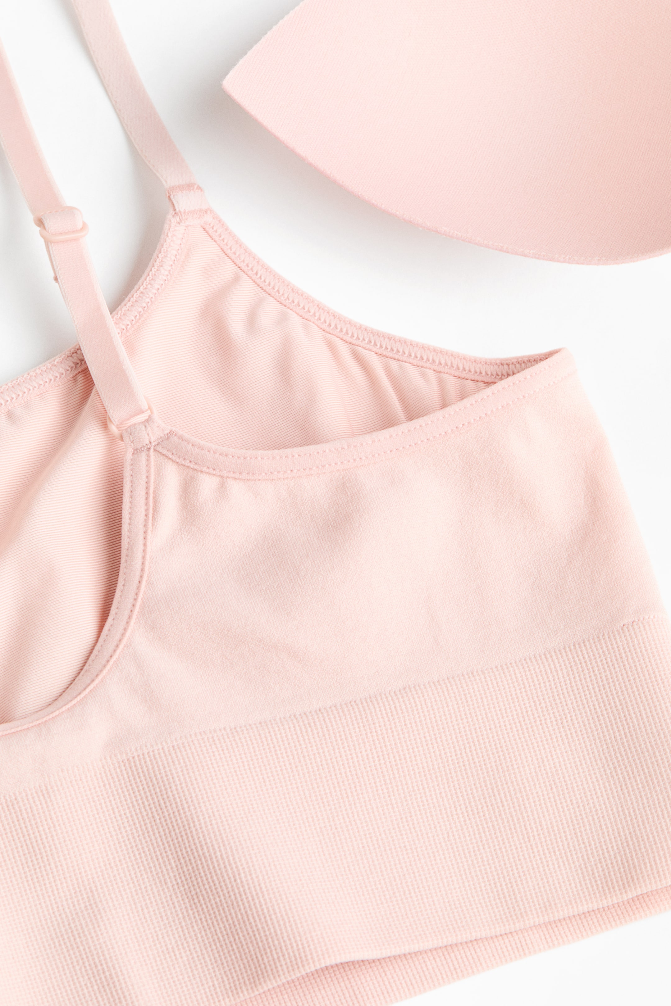 Visualizza l'immagine ingrandita: Un bralette rosa chiaro senza ferretto è esposto, caratterizzato da un corpo anteriore liscio e spalline regolabili. Le coppe sono foderate e un'ampia fascia a coste forma l'orlo, mentre un inserto rimovibile per le coppe abbinato è posizionato nelle vicinanze.