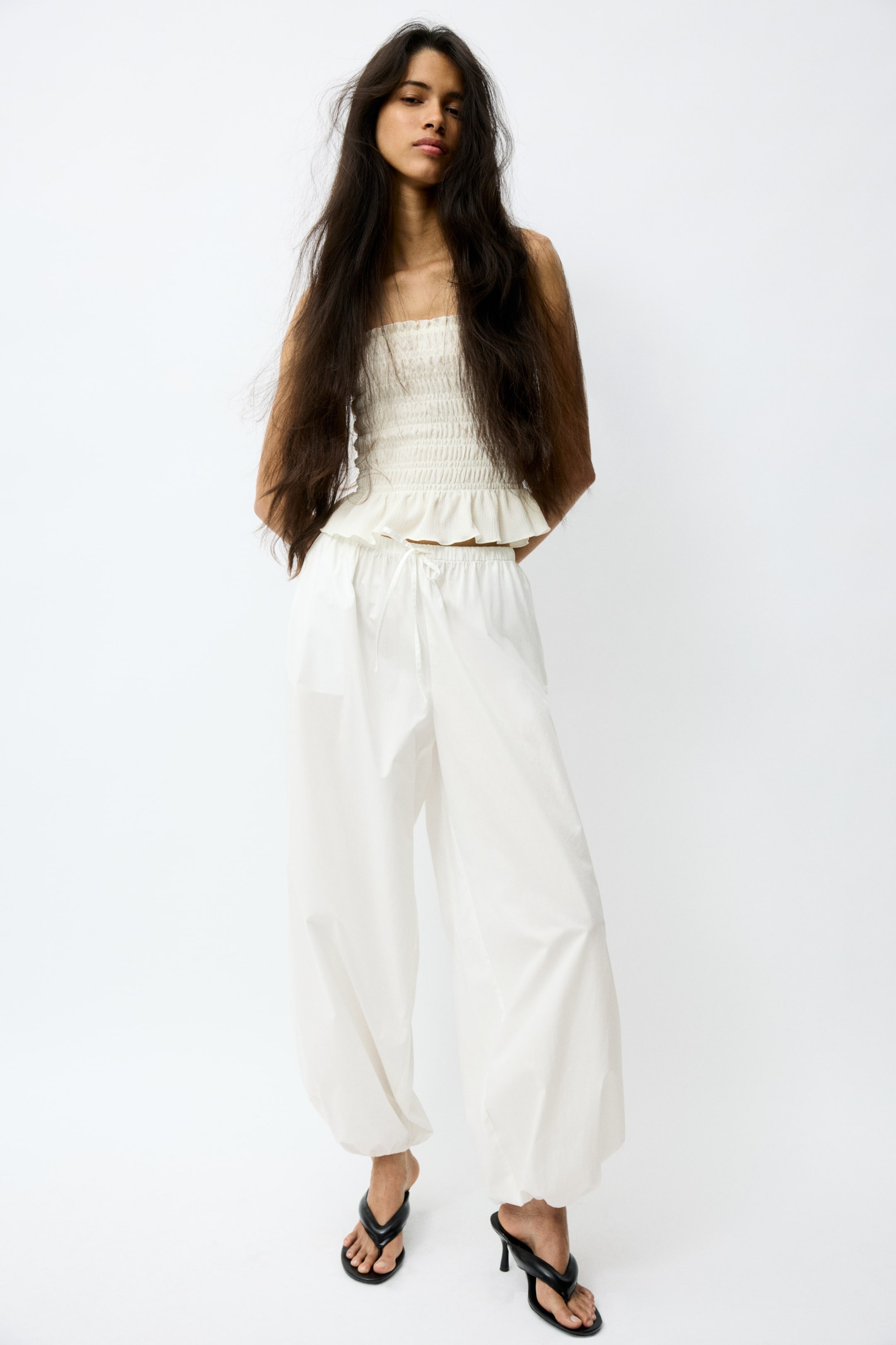 View larger image: An off-white strapless bandeau top features a shirred bodice and a ruffled peplum hem. It is paired with white wide-leg trousers showcasing a drawstring waist and gathered ankles.