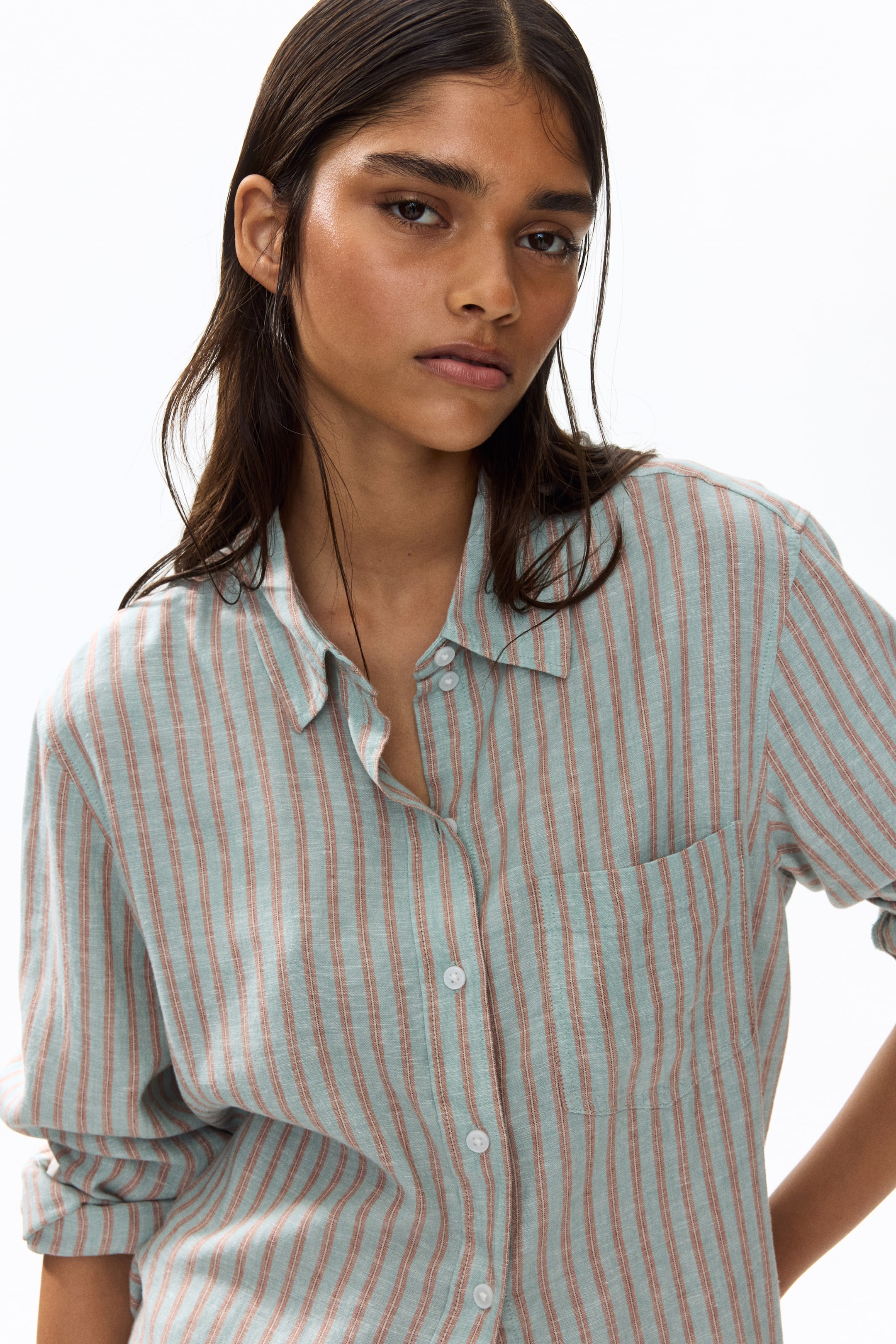 View larger image: The model is donning a loose-fit shirt crafted from a lightweight fabric, featuring vertical stripes in light blue and reddish-brown hues. The shirt showcases a collar, white buttons running down the front, and an open chest pocket. The model has left the top buttons unfastened, with their right hand casually placed on their hip.
