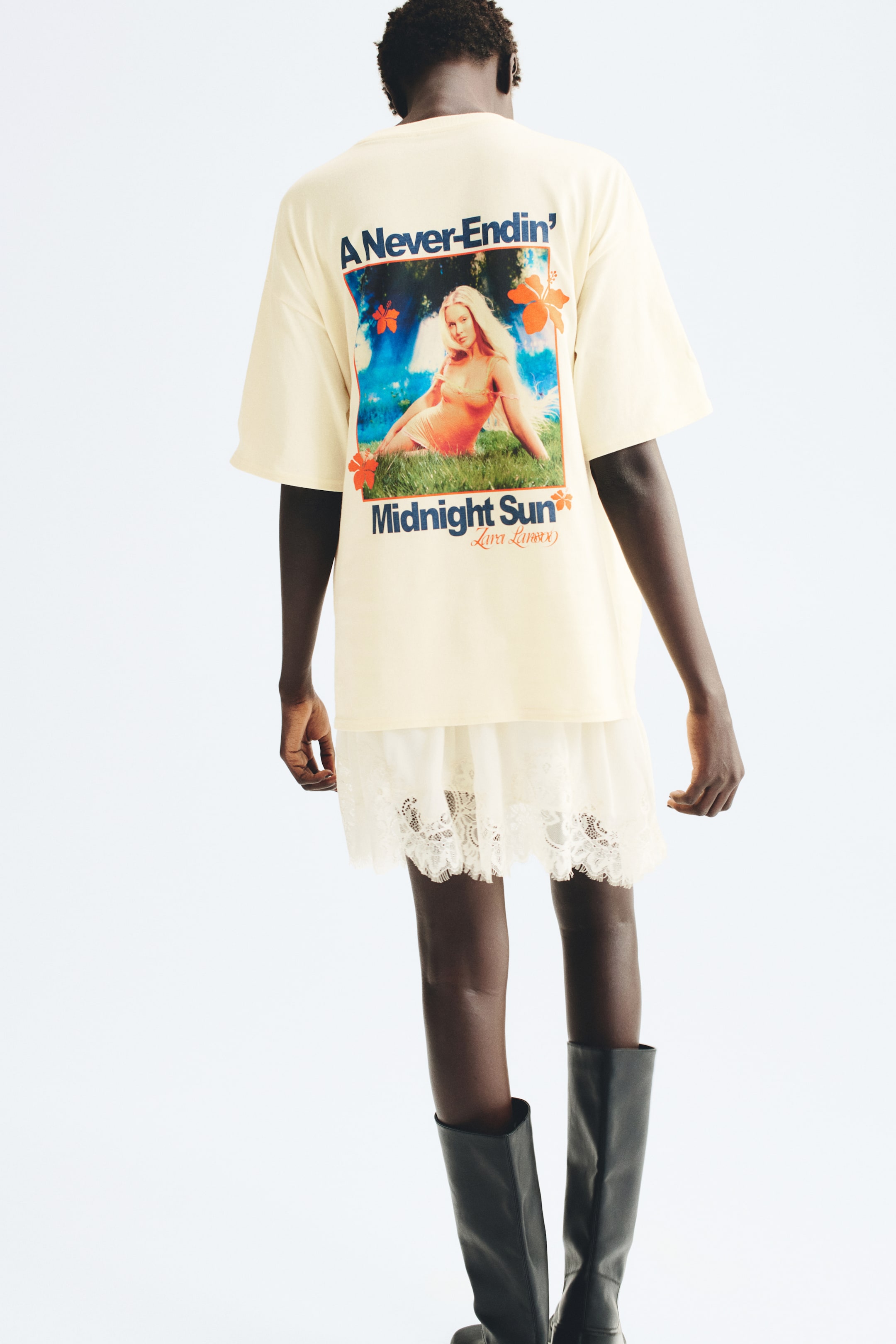 Ver imagem maior: Uma t-shirt creme oversized, com ombros descaídos e uma grande estampagem gráfica colorida nas costas, é usada pela modelo. A t-shirt está sobreposta a uma saia branca com acabamento em renda e combinada com botas pretas altas.