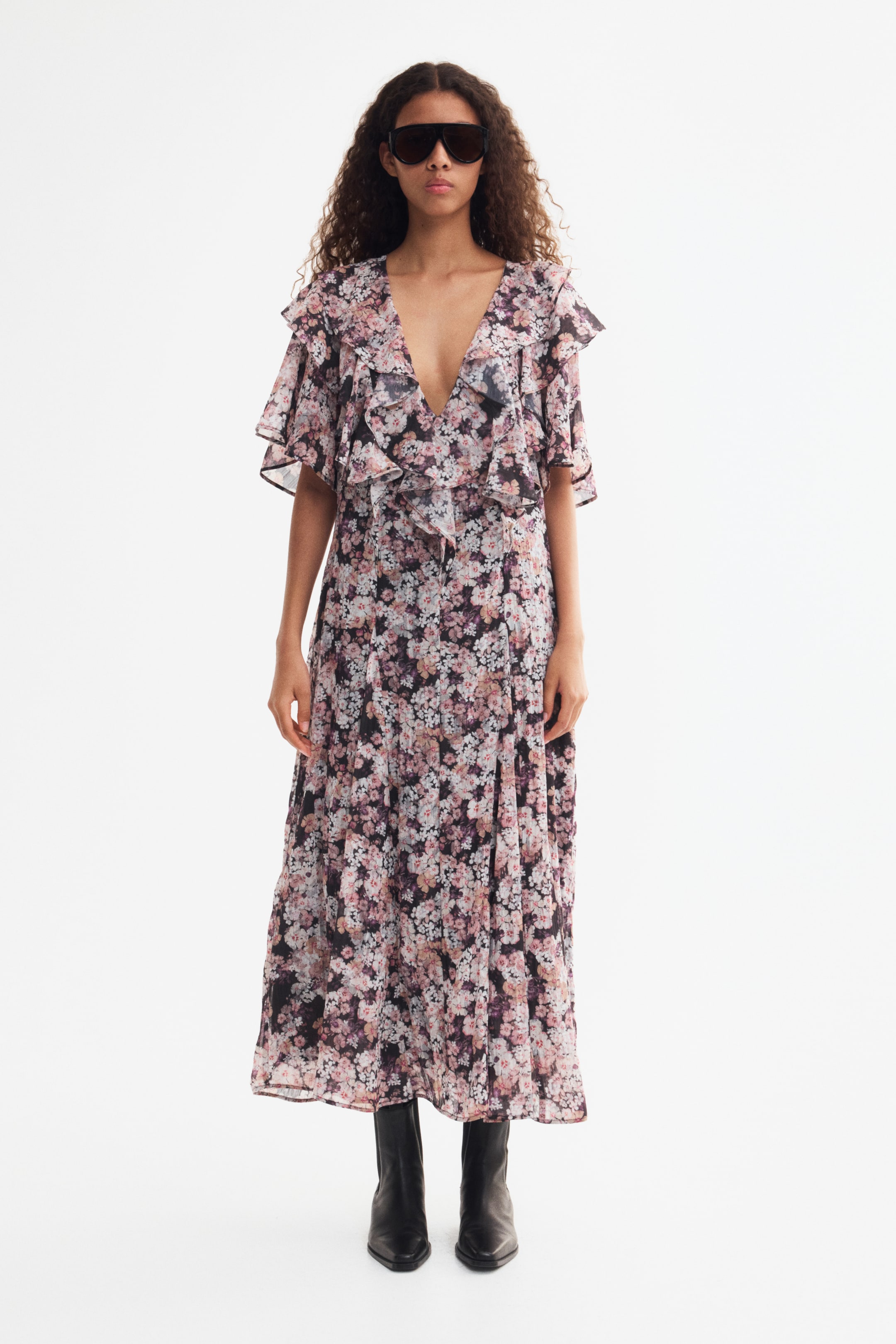 View larger image: A floral-print midi dress featuring a deep V-neckline, ruffled details along the bodice, and short flutter sleeves is worn by the model. The dress is paired with black sunglasses and black leather boots.