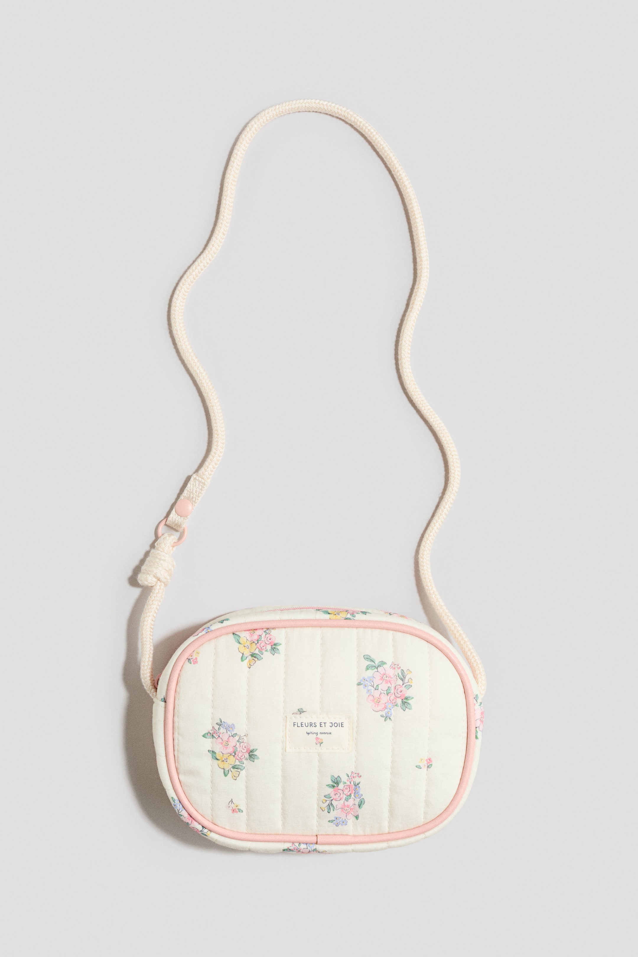 Voir une image plus grande: Un petit sac matelassé présente son DEVANT, avec un tissu blanc cassé parsemé d'un motif floral en rose, jaune et bleu, et affiche une étiquette "FLEURS ET JOIE". Il est conçu avec un passepoil rose le long de sa forme ovale et une fermeture à glissière rose assortie, et une bandoulière en corde couleur crème est fixée au sommet.