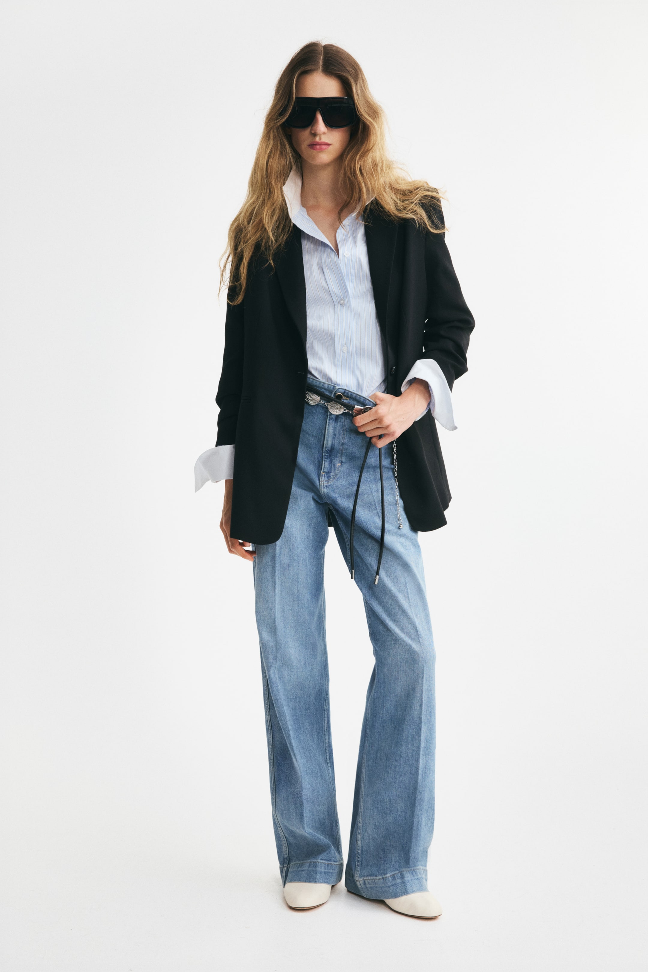View larger image: A long, black single-breasted blazer is shown facing front, featuring notch lapels and structured shoulder pads. The open-front blazer is layered over a light blue striped shirt, with the shirt’s white cuffs folding over the blazer’s three-quarter-length sleeves.
