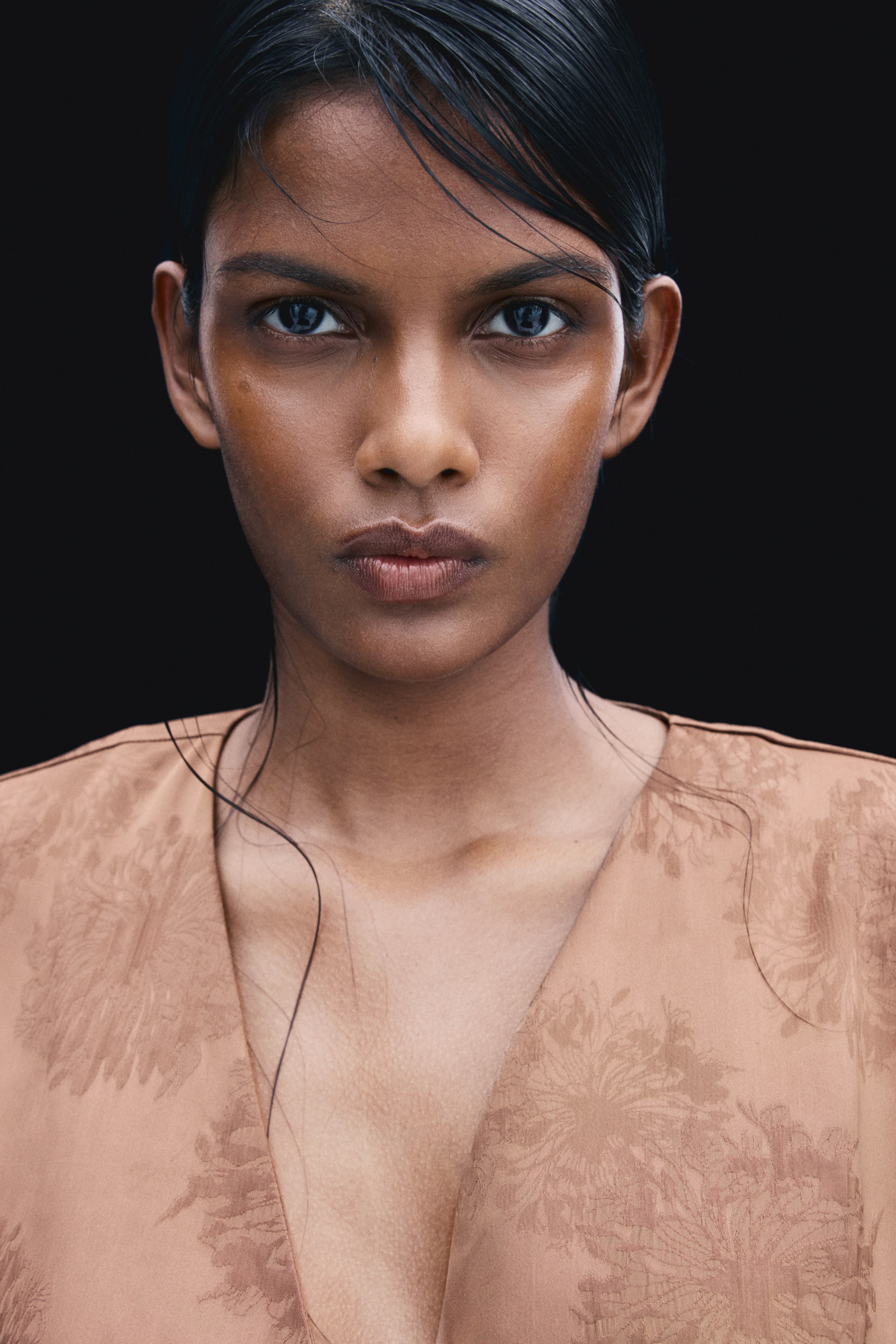 View larger image: The model is wearing a beige jacquard-weave blazer with a subtle floral pattern and a slight sheen. The close-up shot highlights the garment’s deep V-shaped neckline and wrap-front design.
