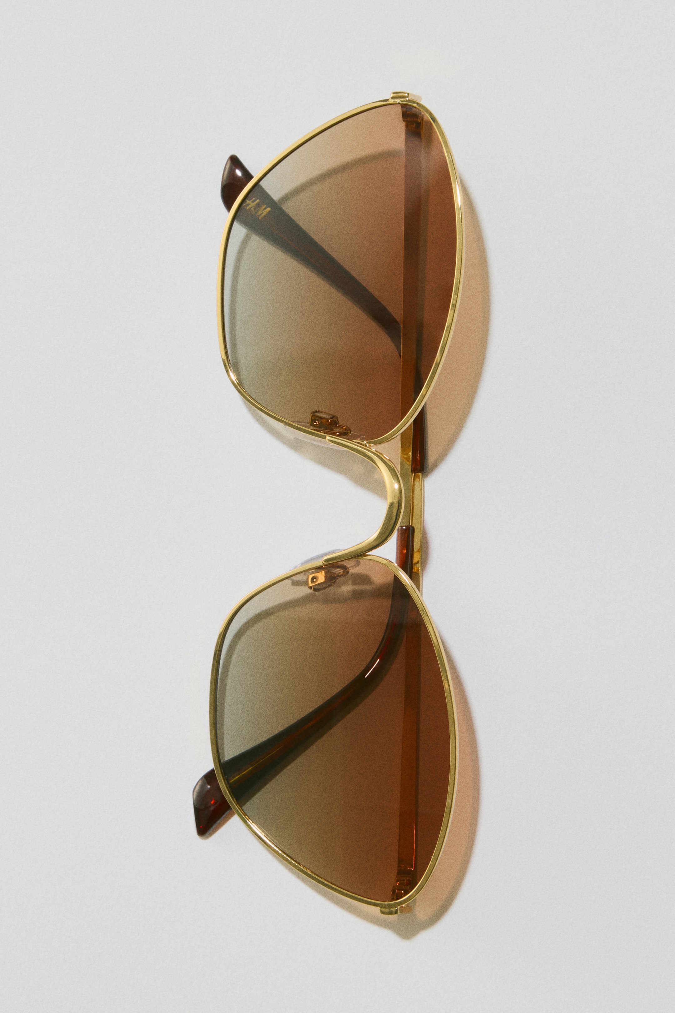 View larger image: Aviator-style sunglasses feature thin gold-toned metal frames with a double bridge. The lenses display a brown gradient tint, darkening from top to bottom. Dark brown temples are visible, with 'H.M' lettering on the left inner temple.