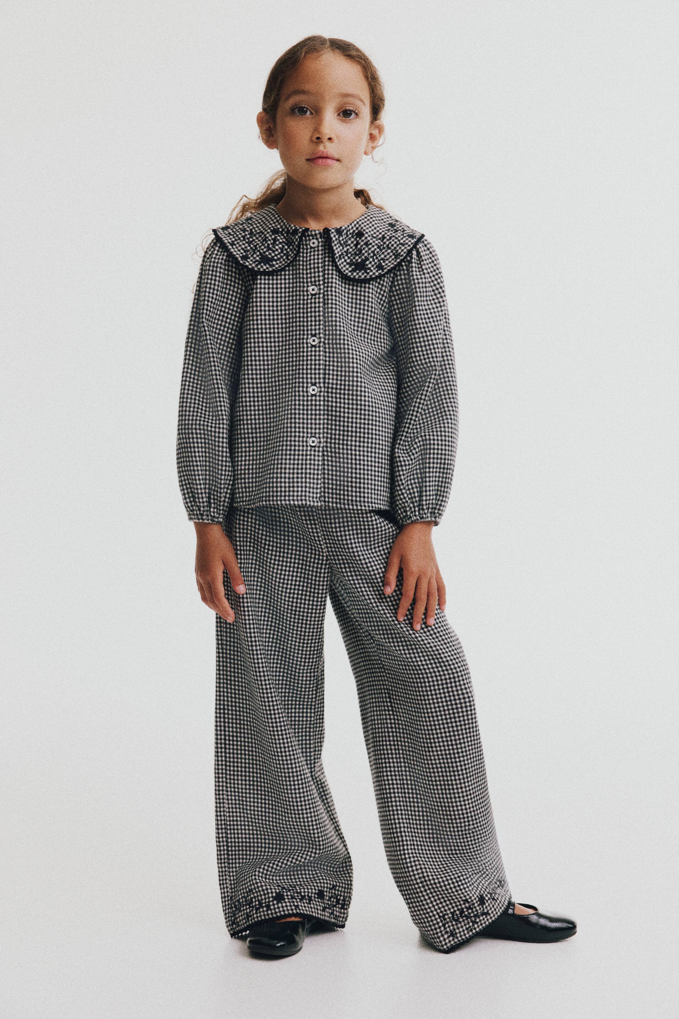 Prikaži veću sliku: Ove pantalone ravnog kroja u crno-belom gingham dezenu prikazane su spreda, sa tamnim cvetnim vezom i delikatnim picot obrubom duž donjih ivica nogavica. Pantalone se nose uz odgovarajuću košulju dugih rukava na kopčanje.