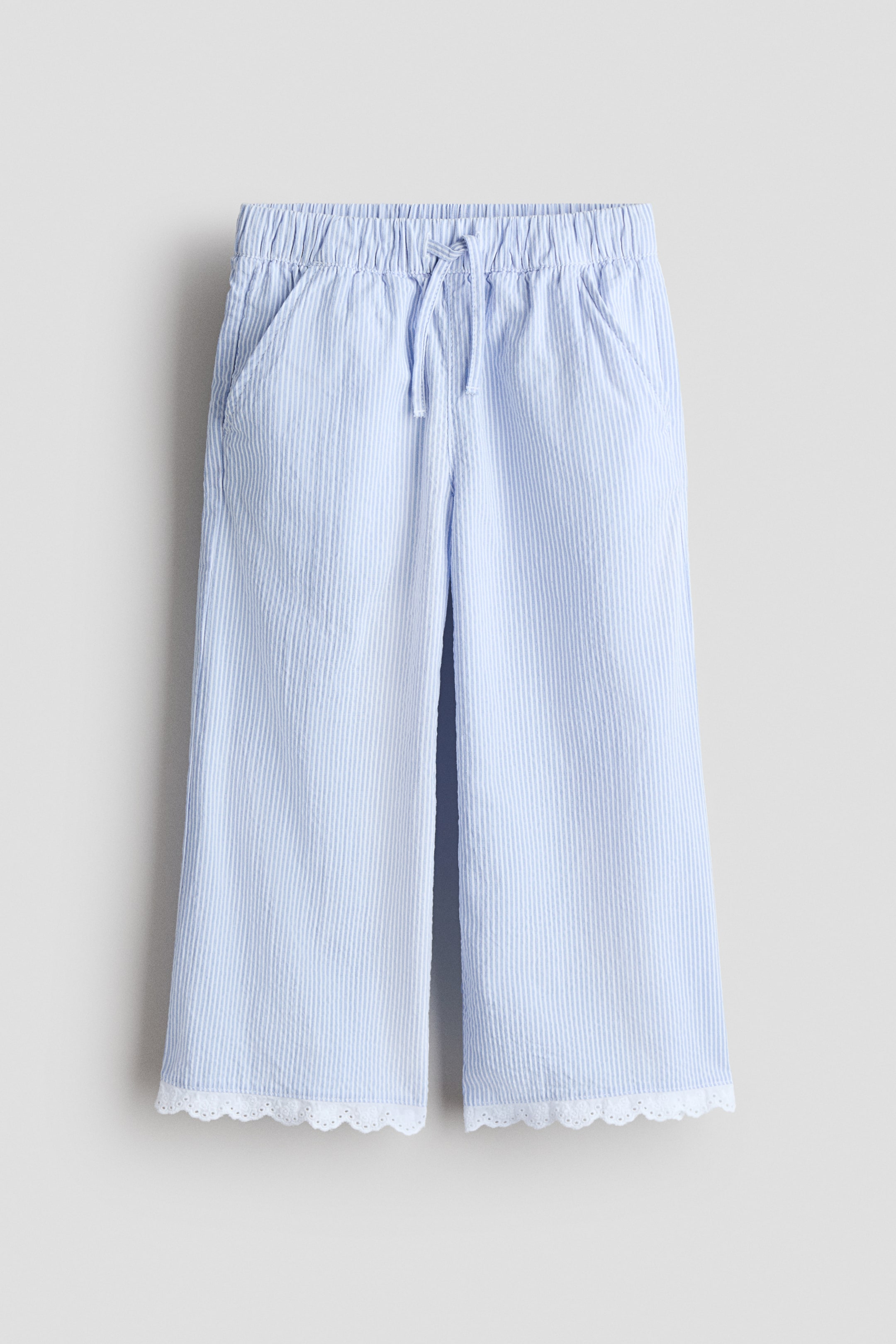 View larger image: These wide-leg trousers showcase a light blue and white seersucker vertical stripe pattern, complete with an elasticized waistband and a drawstring tie at the front. Two side-seam pockets are visible, and a white scalloped lace trim adorns the bottom hems.