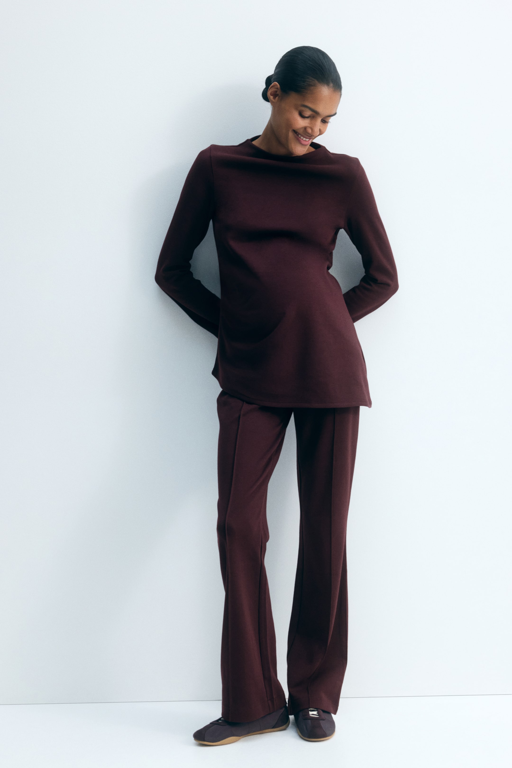 Agrandir l'image: Le mannequin porte un ensemble deux pièces bordeaux foncé, composé d’un haut à manches longues avec col montant et ourlet asymétrique. Le pantalon assorti présente une taille haute avec des plis cousus sur le devant des jambes évasées.