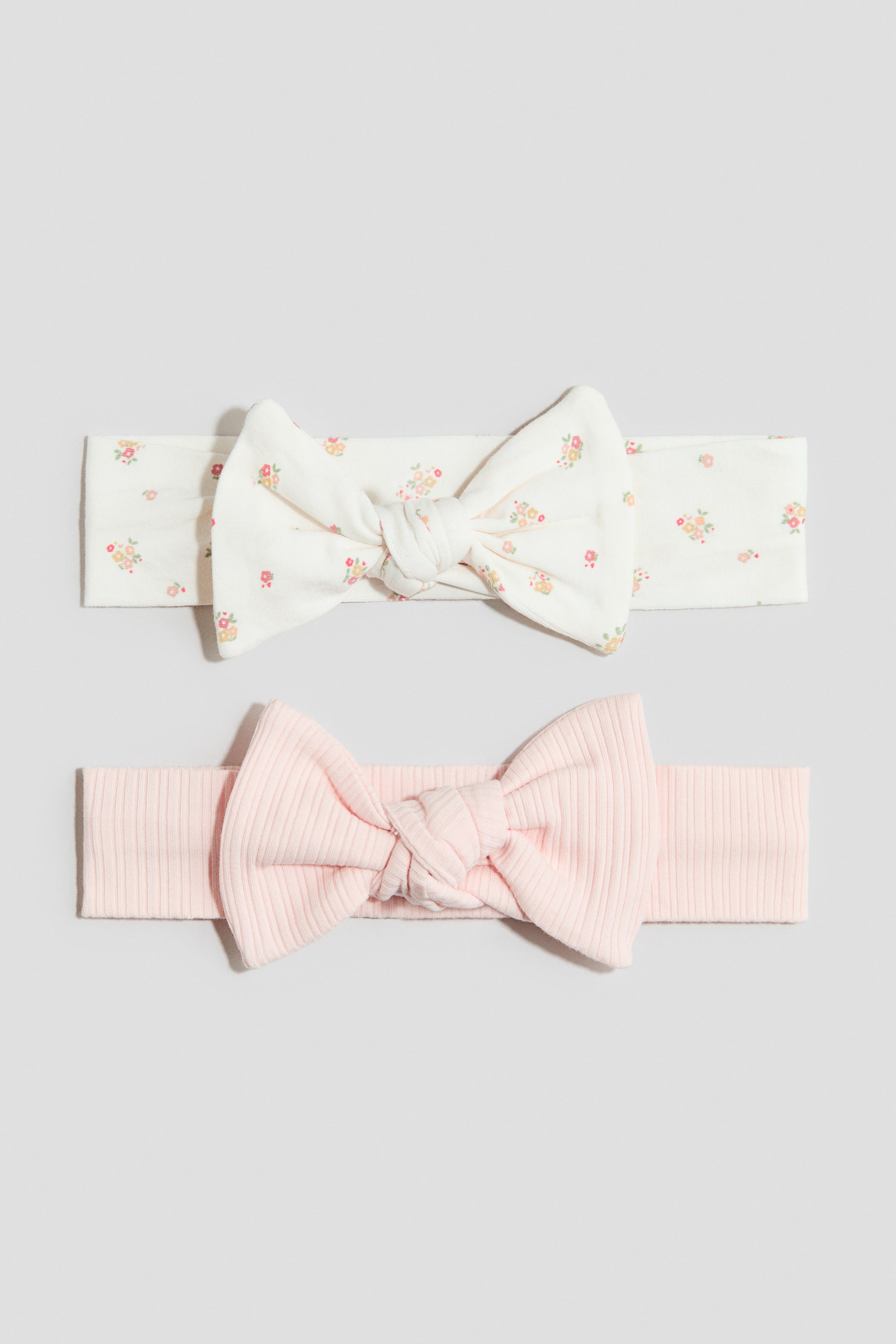 이미지 확대 보기: 부드러운 코튼 저지 소재의 헤어밴드 2개가 정면에서 보이도록 배치되어 있으며, 각각 리본 장식이 있습니다. 한쪽은 작은 플로럴 프린트가 들어간 오프화이트 색상이고, 다른 한쪽은 골지 텍스처의 연핑크 색상입니다.