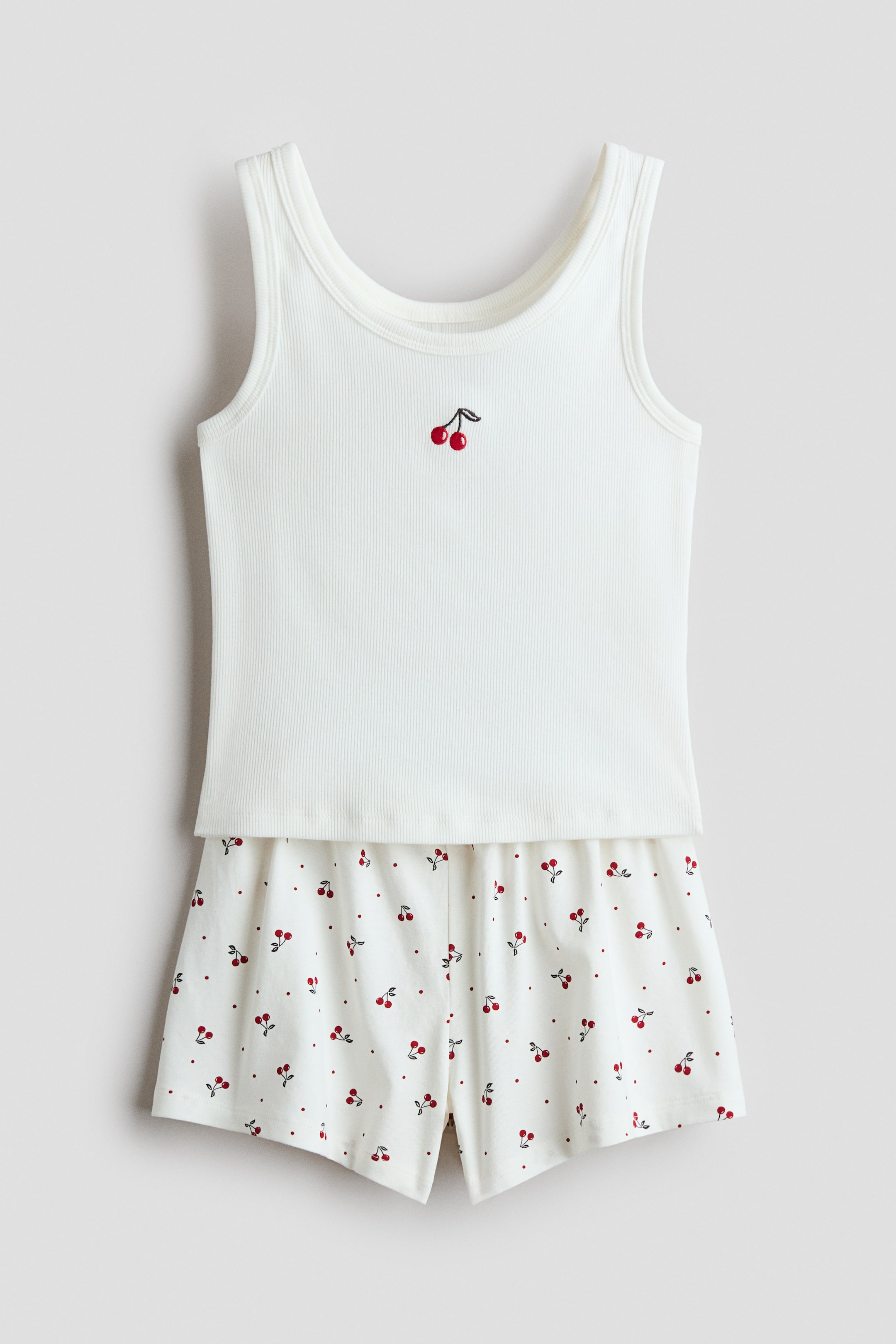View larger image: A set of pyjamas featuring a white ribbed vest top paired with matching white shorts, presented facing front. The top boasts a small embroidered cherry motif on the front, with binding around the neckline and armholes, while the shorts display an all-over cherry and dot print with an elasticated waist.