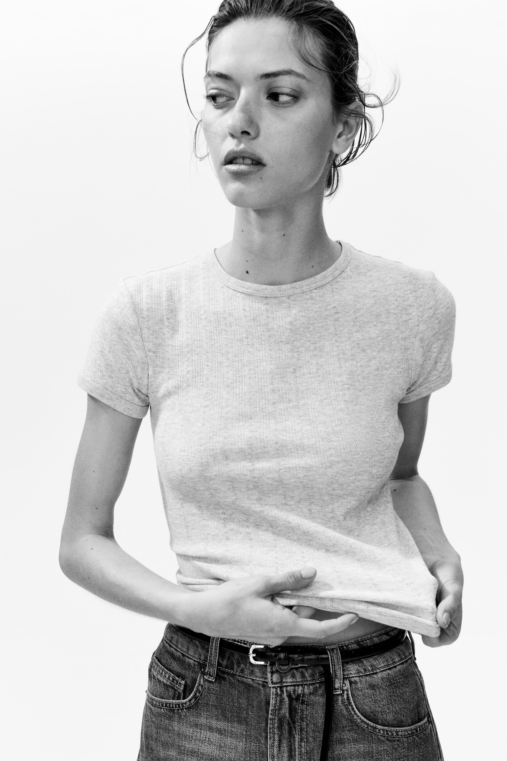 View larger image: A light-colored ribbed knit t-shirt featuring a crew neck and short sleeves, adorned with small eyelet details creating vertical patterns on the fabric. The bottom hem is held up by hands.
