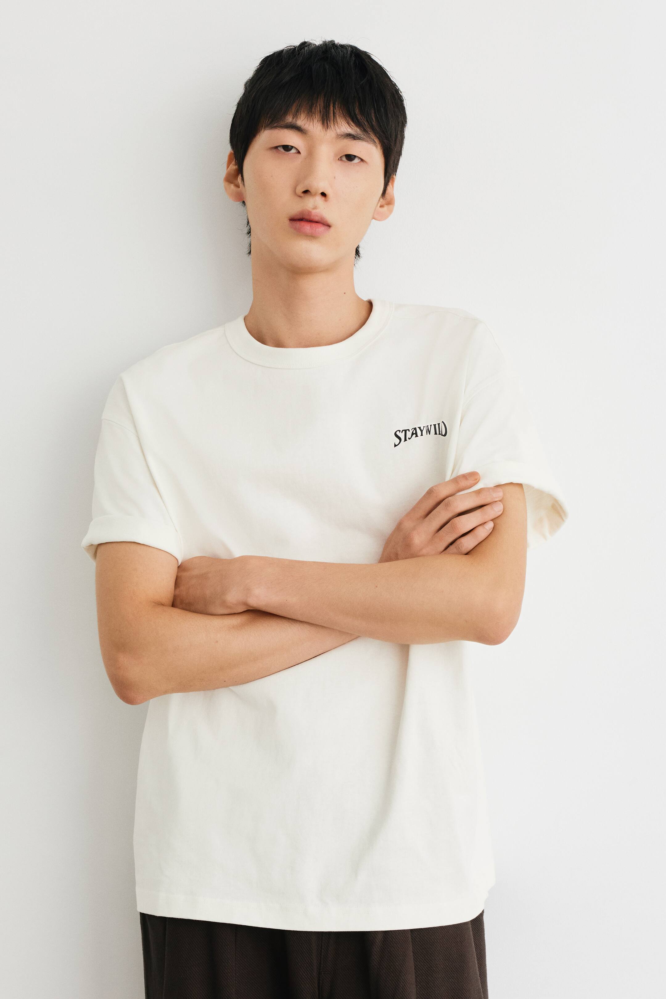 View larger image: An off-white, loose-fit T-shirt in heavyweight cotton jersey is worn by the model, who is standing with arms crossed. The T-shirt features a round, rib-trimmed neckline, short sleeves with rolled cuffs, and a small black graphic print reading "STAY WILD" on the left chest.