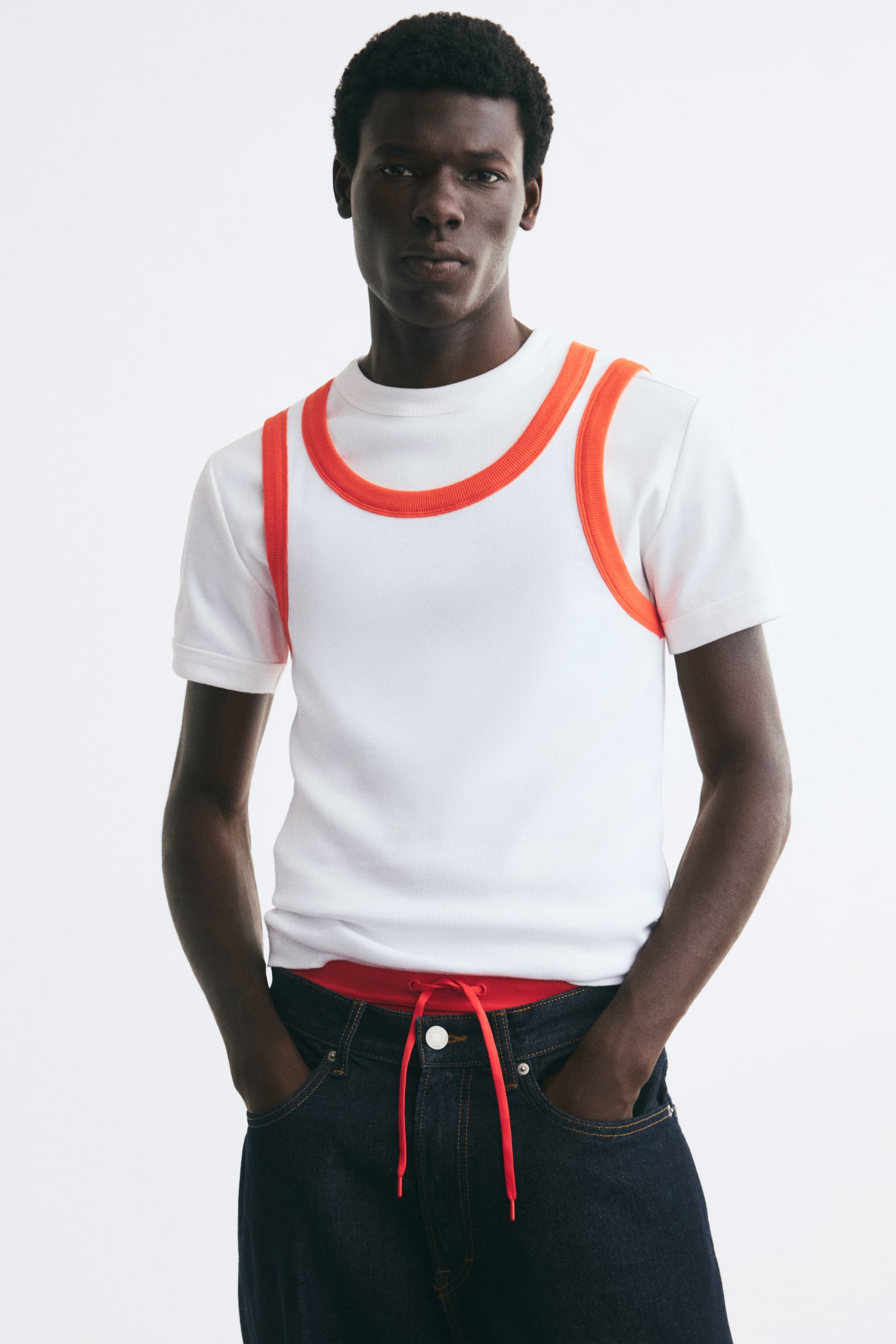 A white ribbed jersey tank top with bright orange contrast trim along the scoop neck and armholes is worn by the model. The slim-fit tank is layered over a short-sleeved white T-shirt and paired with dark denim jeans. - 1