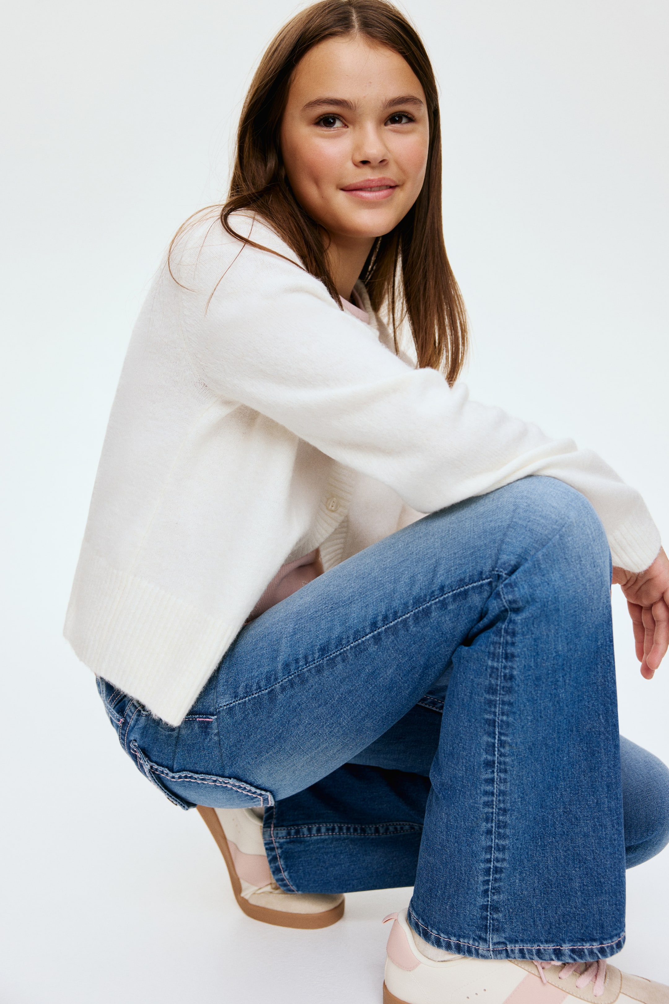 View larger image: A white button-front cardigan, featuring a fuzzy texture and ribbed cuffs and hem, is being worn over a pink top. It is paired with blue bootcut jeans, seen from a side-back angle, which are showing pink stitching details on the back pockets and a slight flare at the ankles.