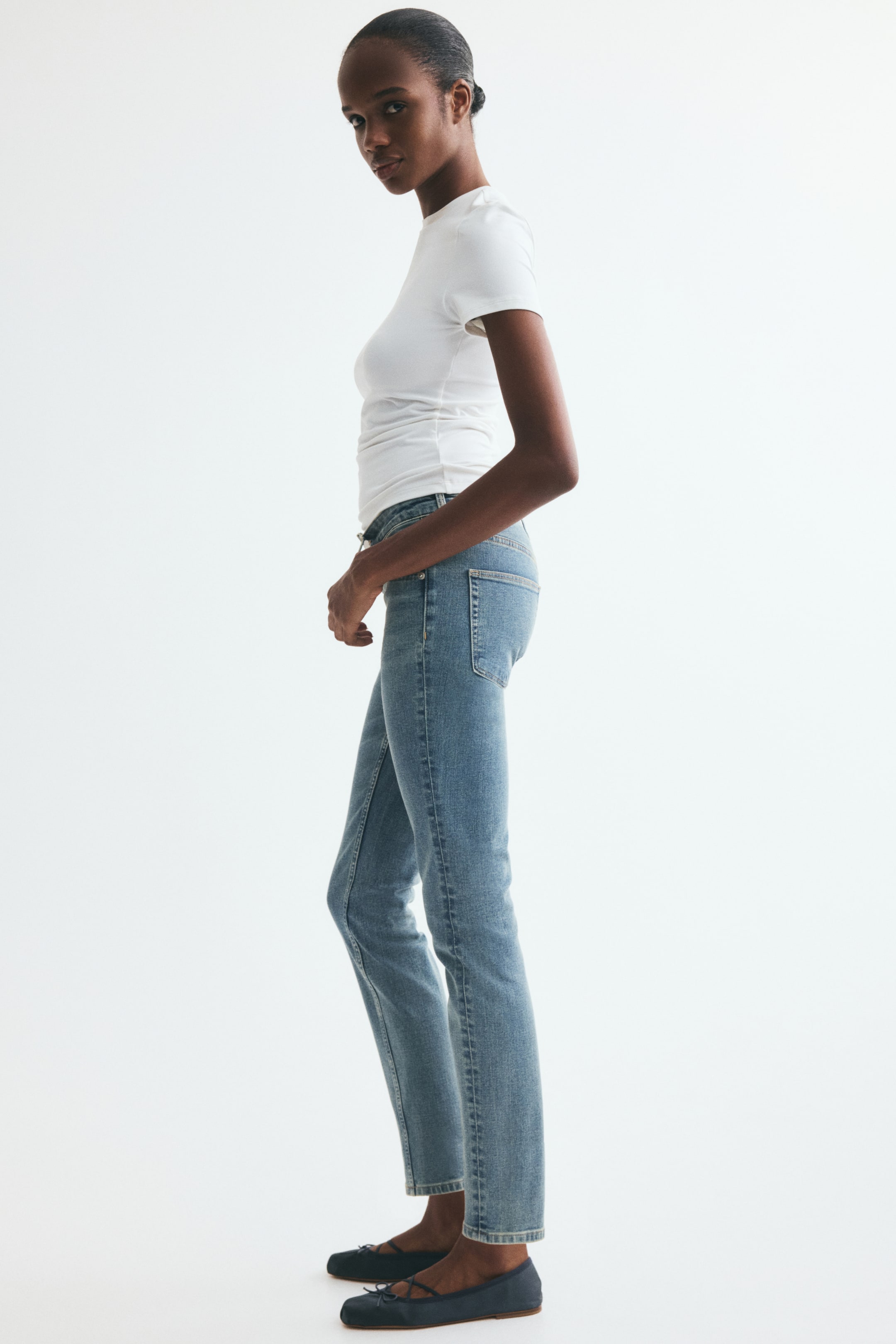 View larger image: The model is wearing a white, short-sleeved fitted T-shirt with a round neckline and side ruching, light blue straight-leg jeans, and dark blue ballet flats with a round toe and criss-cross ties.