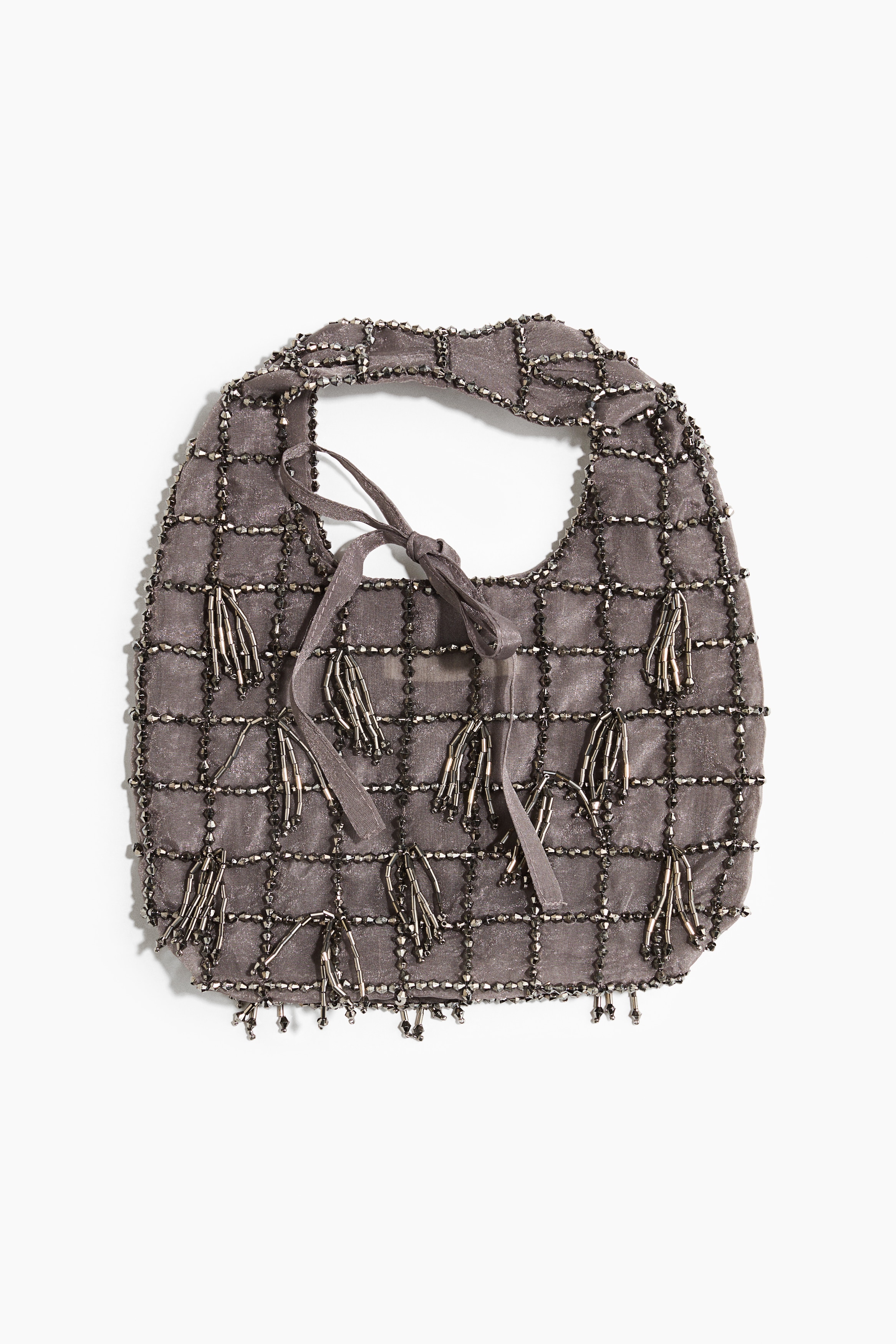 Un petit sac à main en organza gris transparent, orné d’un quadrillage de perles sombres et de pampilles suspendues, est porté par le mannequin, qui le tient par sa poignée supérieure. Le sac à main est associé à un pantalon noir et à des escarpins noirs à bout pointu.