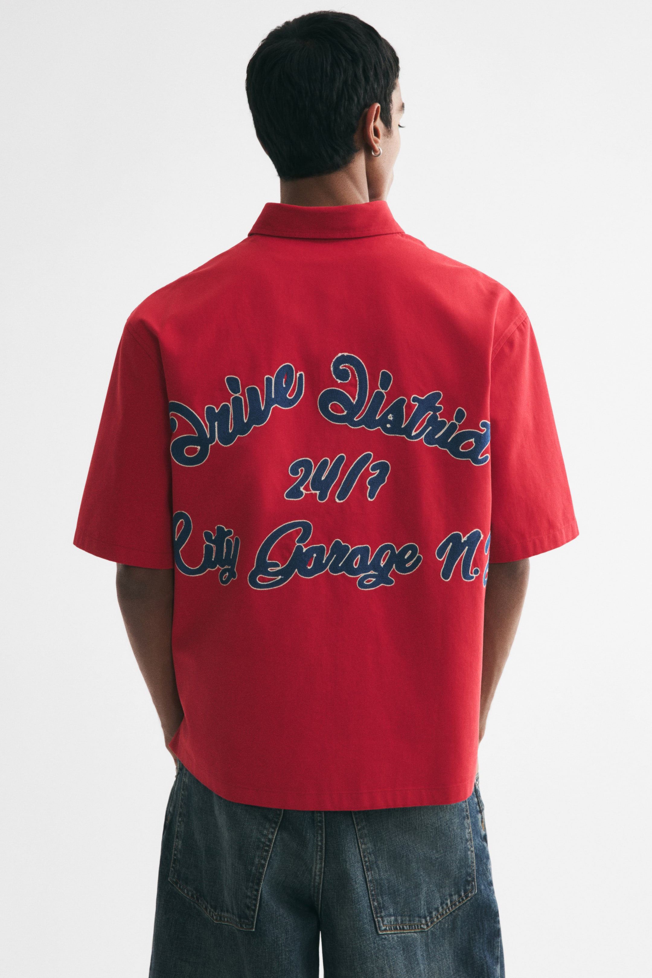 View larger image: A red, short-sleeved collared shirt with a large blue embroidered text motif across the back is being worn by the model. The model is standing with their back to the camera, showcasing the relaxed-fit shirt styled with blue denim jeans.